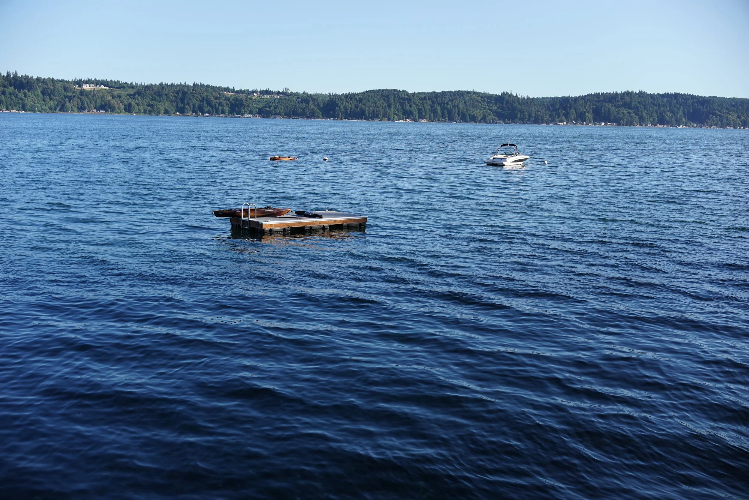 A body of water with a small dock, a kayak, and a motorboat, with a tree-lined shoreline and hills in the background under a clear blue sky.