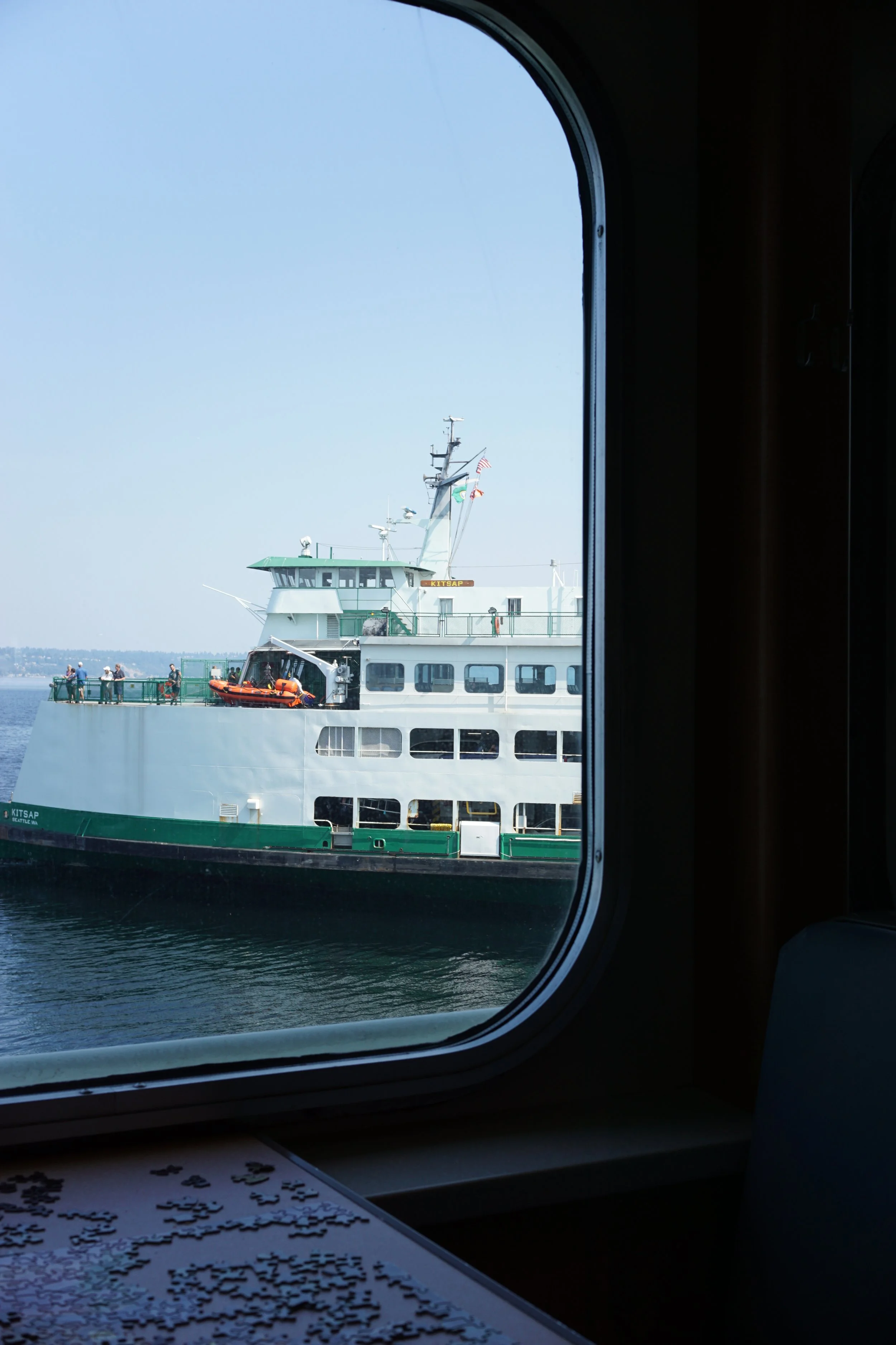 View of a ferry boat through a window on a boat or ferry, with people standing on the deck and ships visible in the water.