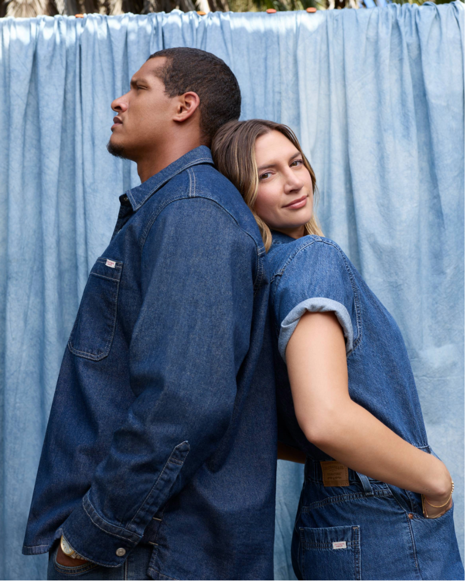 Man and woman standing back-to-back in front of a blue curtain, both wearing denim jeans and shirts, with the woman smiling slightly at the camera.