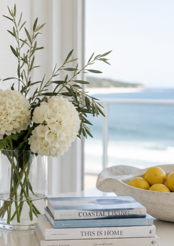 A vase of white hydrangea flowers and green leaves on a table with a white bowl of lemons, a stack of books, and a view of the ocean through a window.