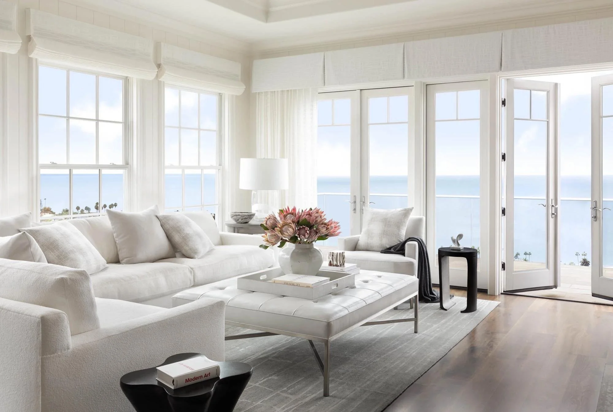 Living room with white sectional sofa, coffee table, pink flowers, large windows with ocean view, glass doors, side table with sculpture, and a modern black side table with books.