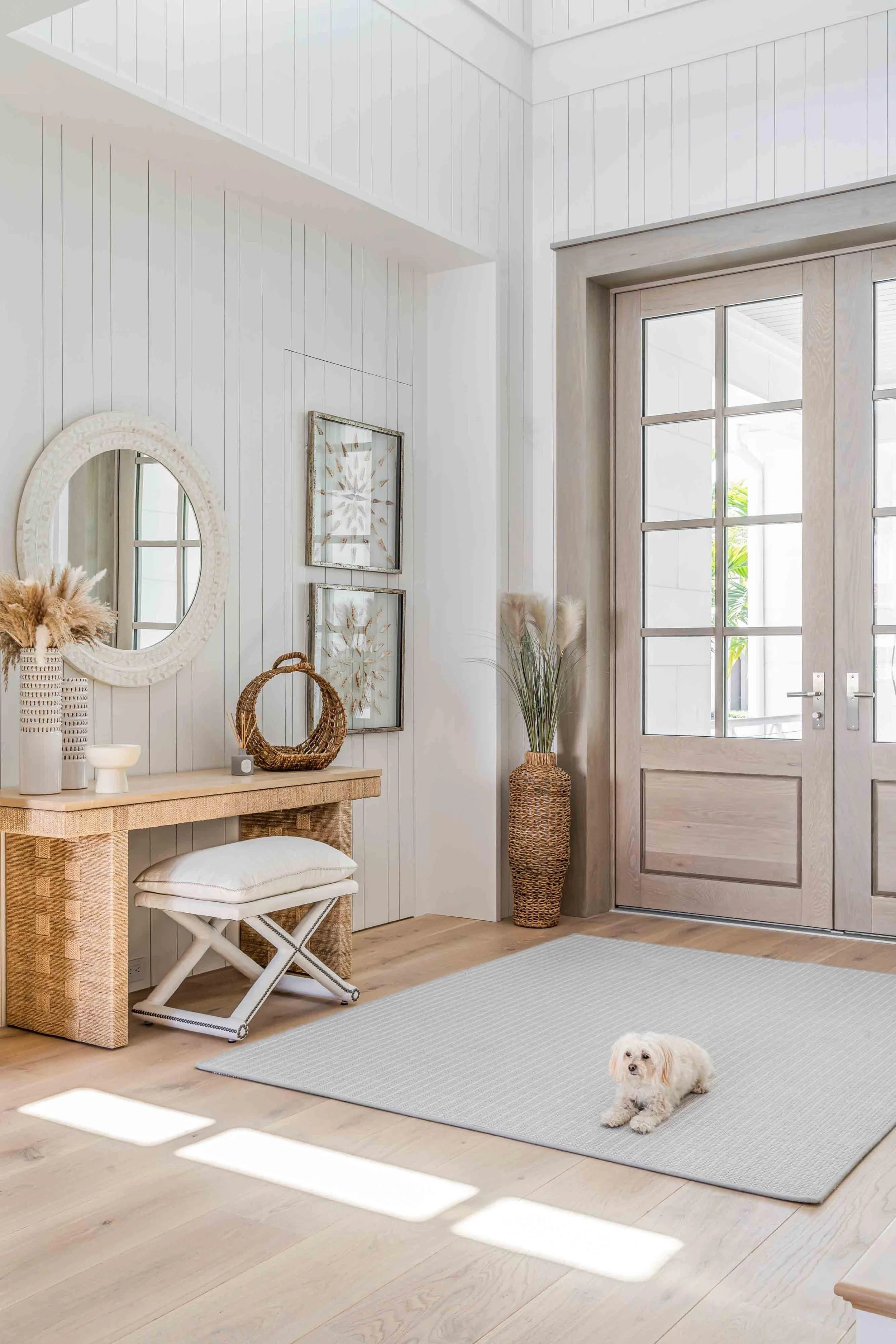 A cozy, Scandinavian-style entryway with a large windowed door, light wood flooring, and white paneled walls. Decor includes a round mirror, framed botanical prints, wicker and ceramic baskets, a small stool with a cushion, a tall wicker vase with pampas grass, and a small white dog lying on a light gray rug.