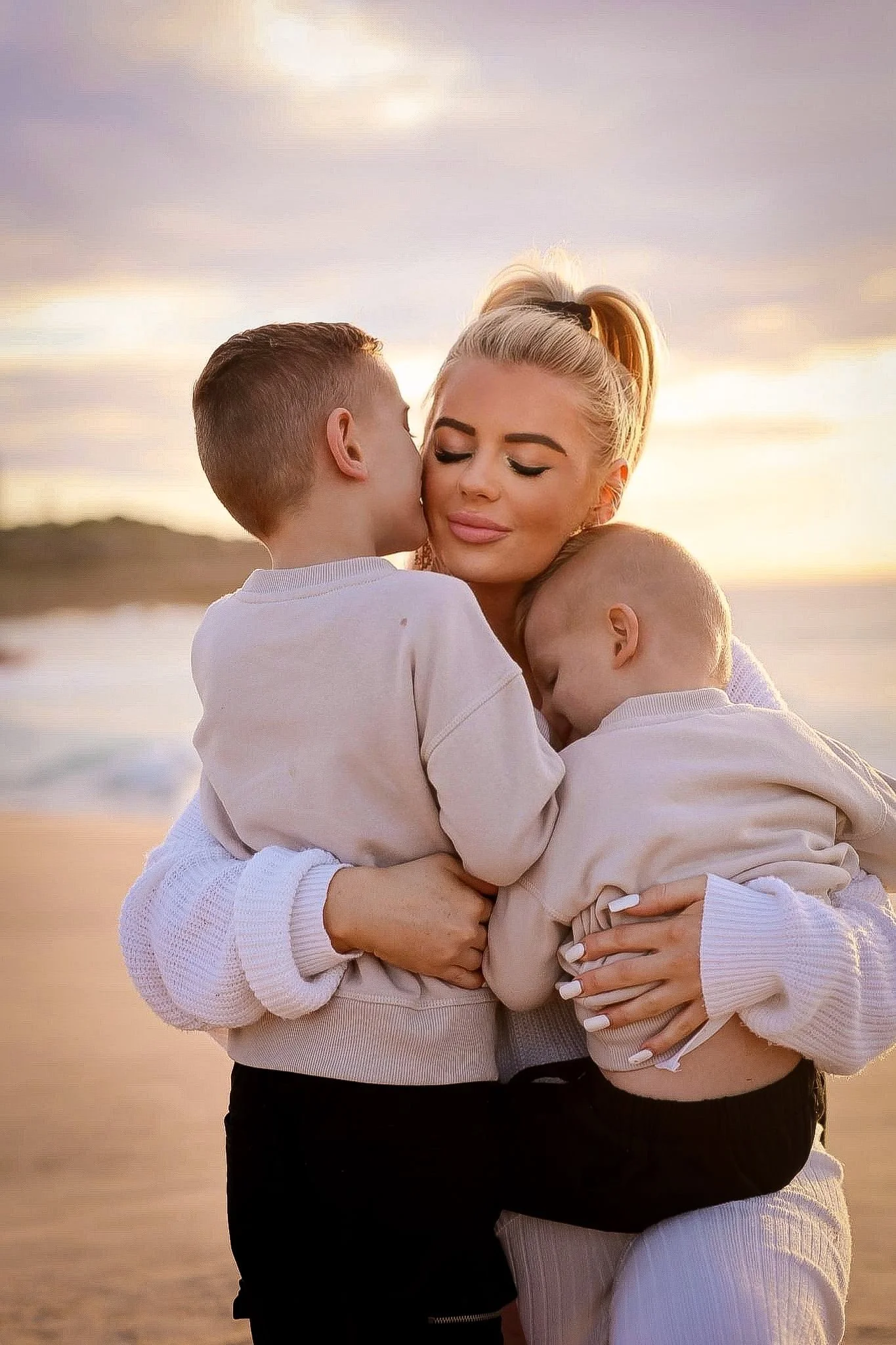 A woman and two children embracing and sharing a kiss on the cheek during sunset at the beach.