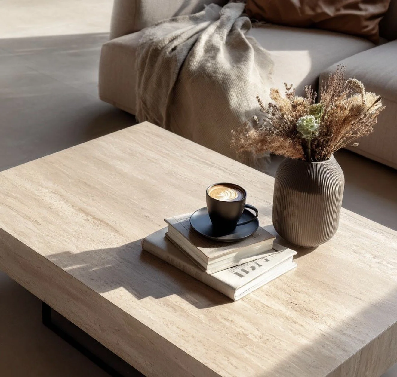 A living room coffee table with a black coffee cup on a saucer, a stack of books, and a beige vase with dried flowers, surrounded by a beige sofa with cushions and a blanket.