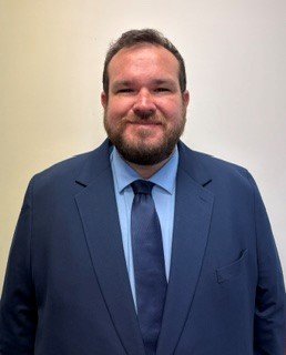 A man wearing a dark blue suit, light blue shirt, and dark tie standing against a plain background.