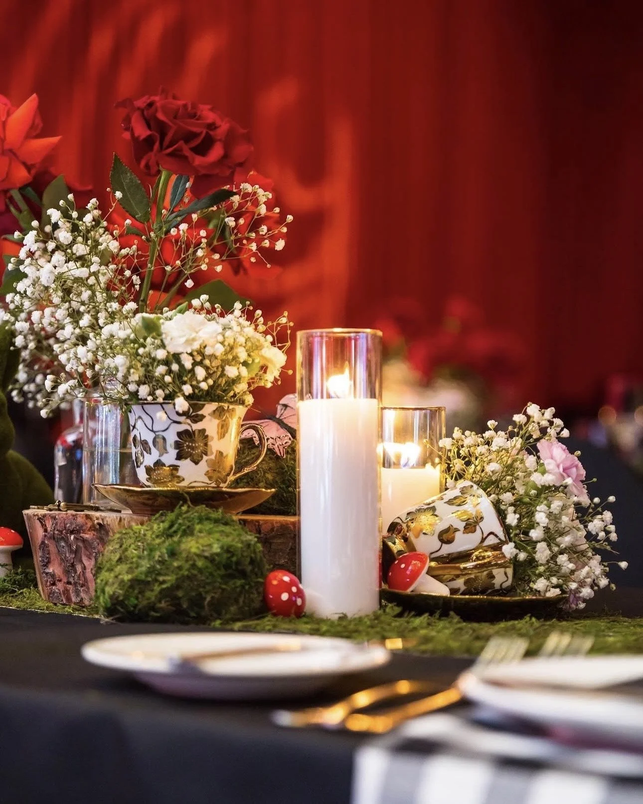 Centerpiece with red and white flowers, lit candles in glass holders, and ceramic mugs with floral designs on a decorated table.