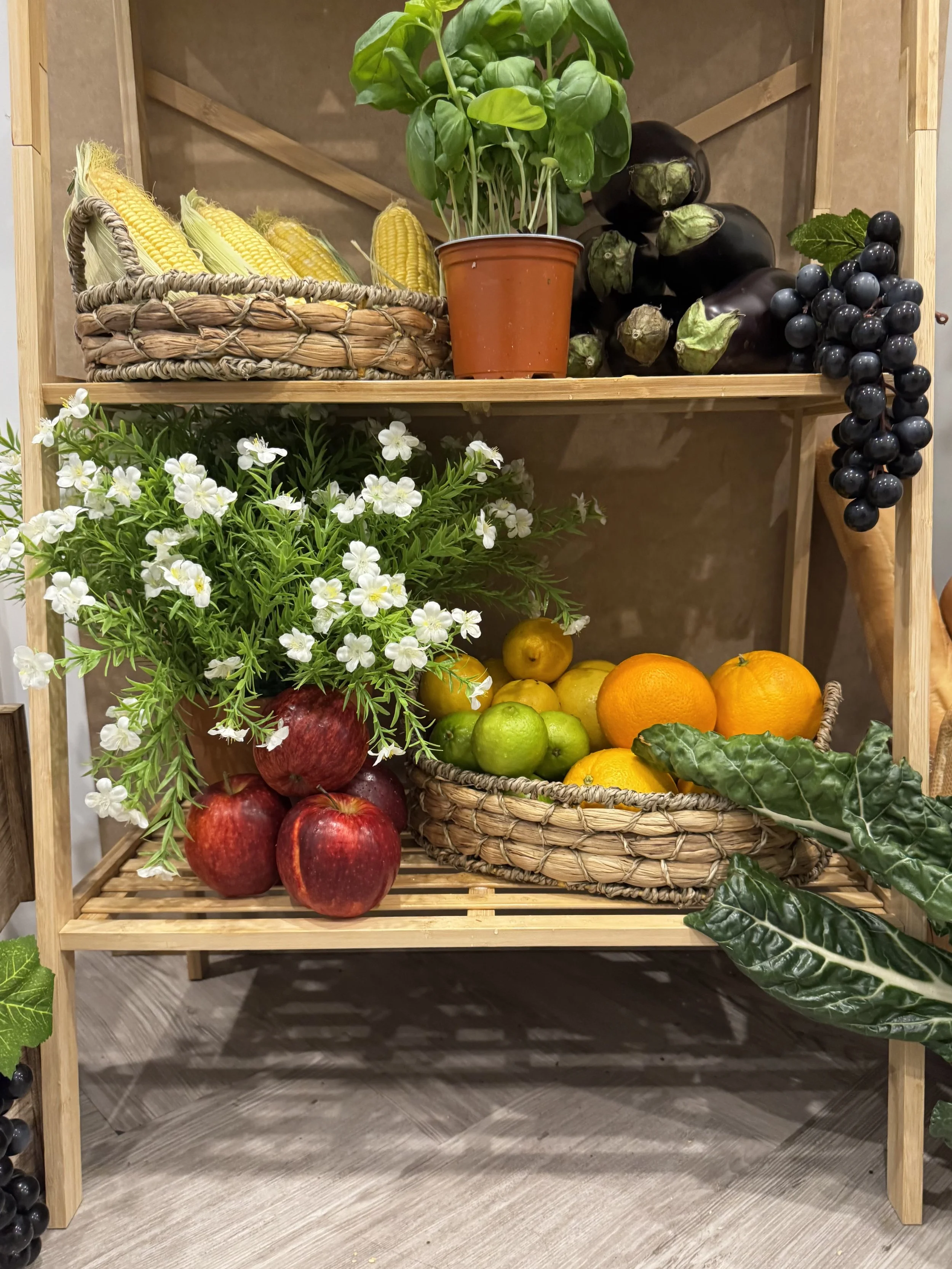 A wooden shelf with fresh produce and plants. Top shelf has corn, basil, eggplants, and grapes. Middle shelf has white flowers, apples, oranges, and a basket of citrus fruits. Bottom shelf is empty, showing a gray wooden floor.