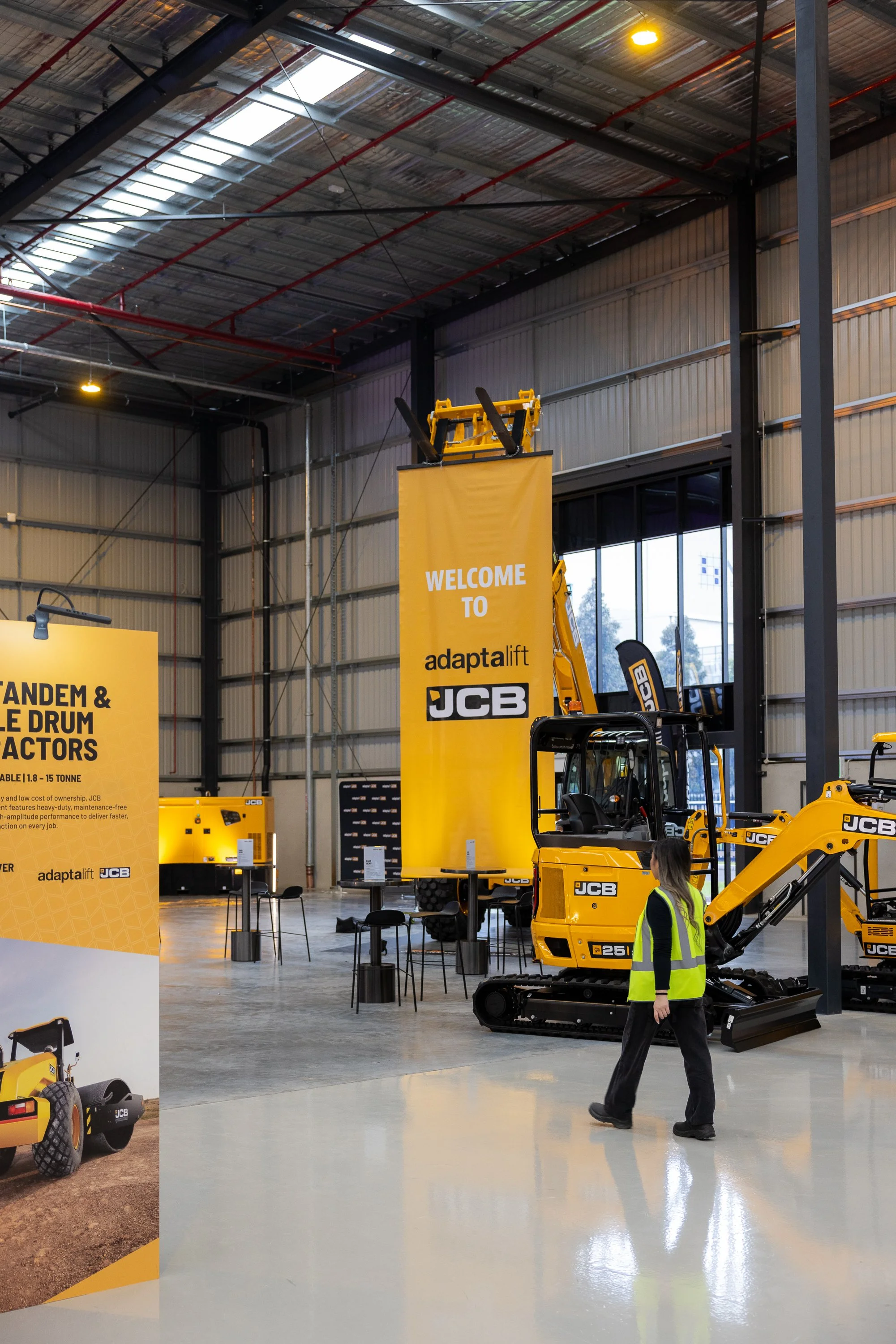 Indoor exhibition space displaying JCB construction equipment, including a yellow mini excavator, with banners and signage for 'adaptalift' and 'JCB'. A woman wearing a yellow safety vest walks past the equipment.