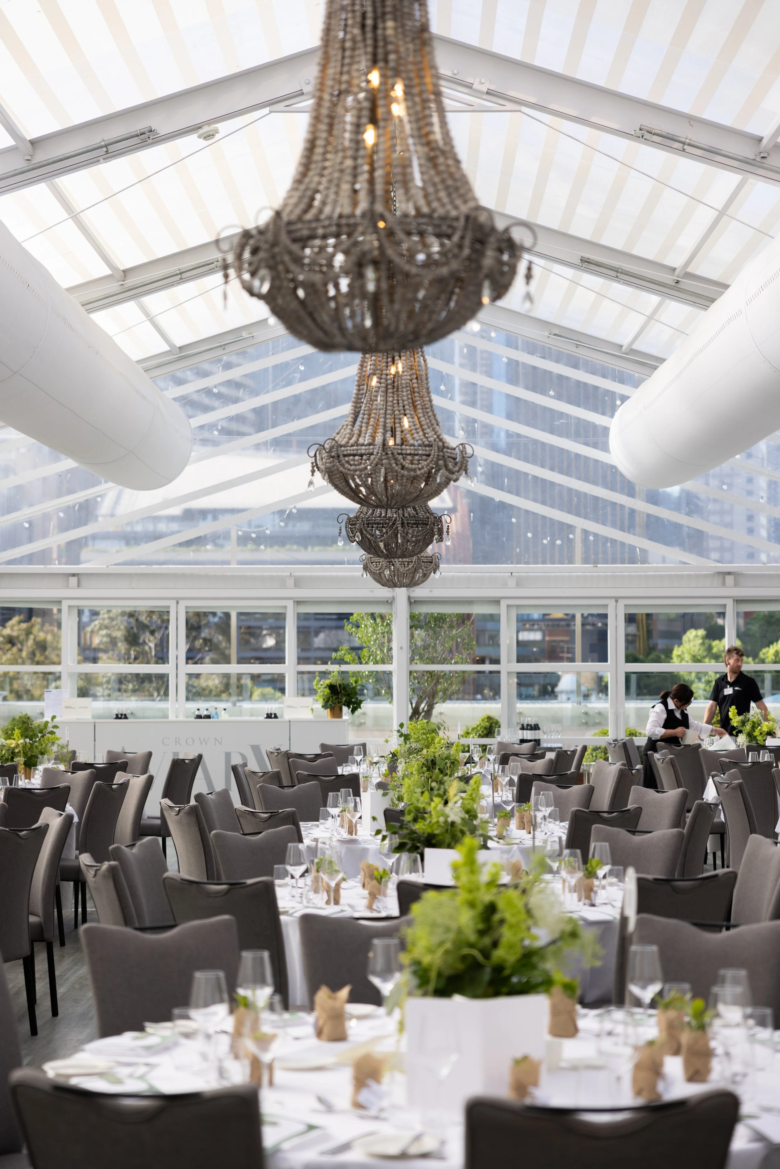 Elegant indoor event space with large chandeliers hanging from a high glass ceiling, set with round tables, gray chairs, and green floral centerpieces, with waitstaff preparing in the background.