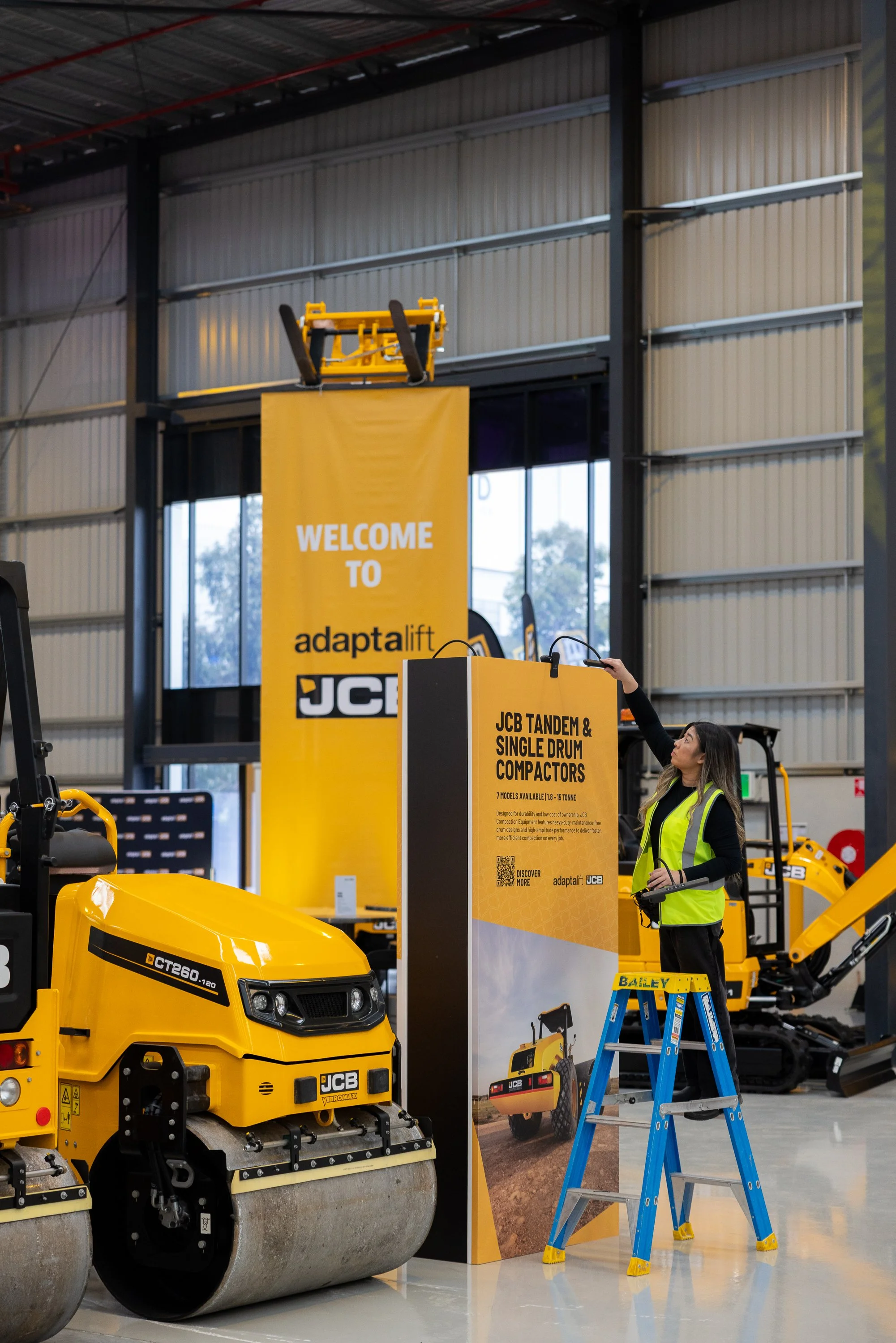 Indoor exhibition of construction equipment, featuring a yellow JCB compact roller and a yellow JCB mini excavator, with promotional banners and a woman in a yellow safety vest standing on a blue ladder.
