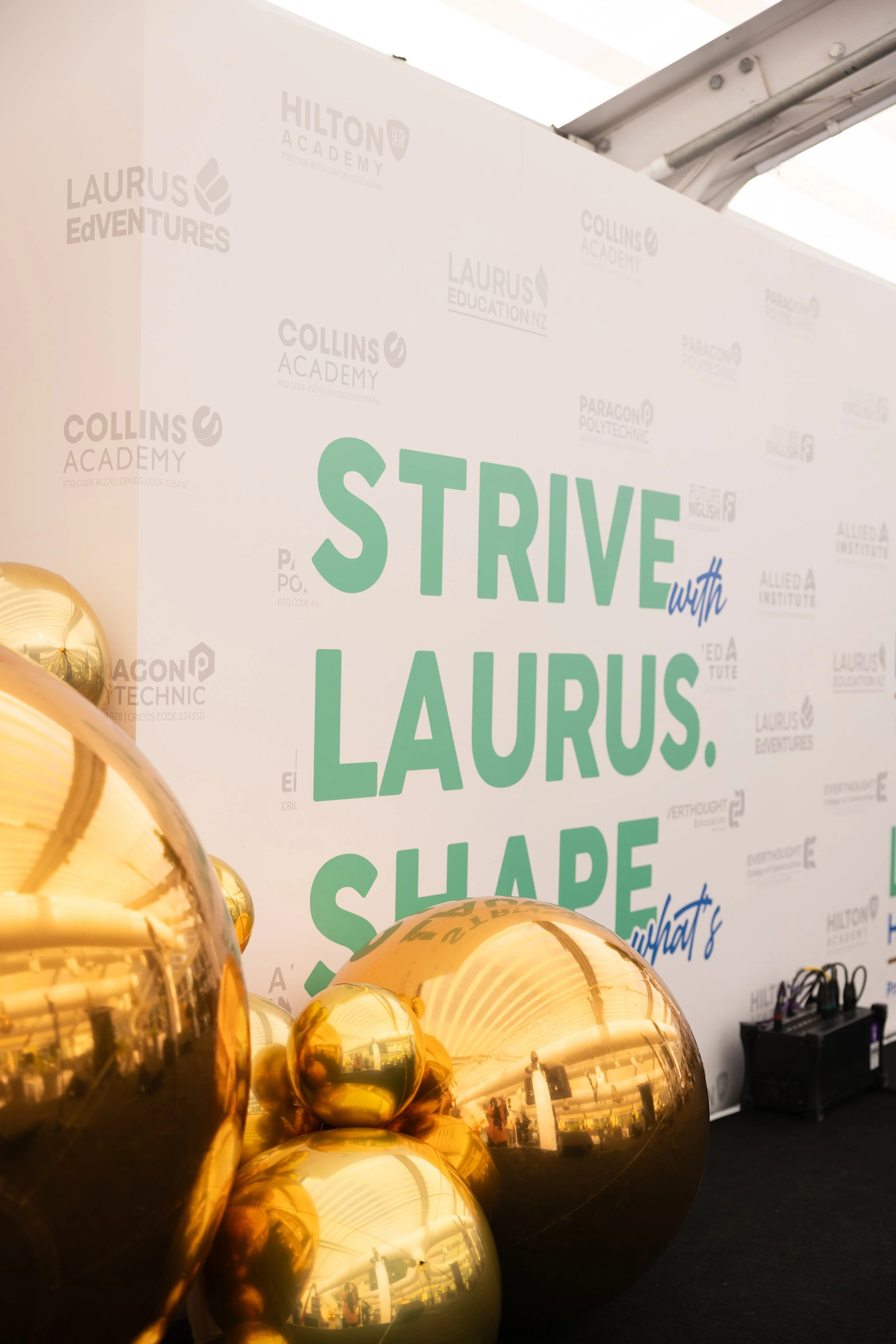 Gold balloons in the foreground with a white backdrop displaying various educational institution logos and the phrase 'Strive with LAURUS' in green and blue text.