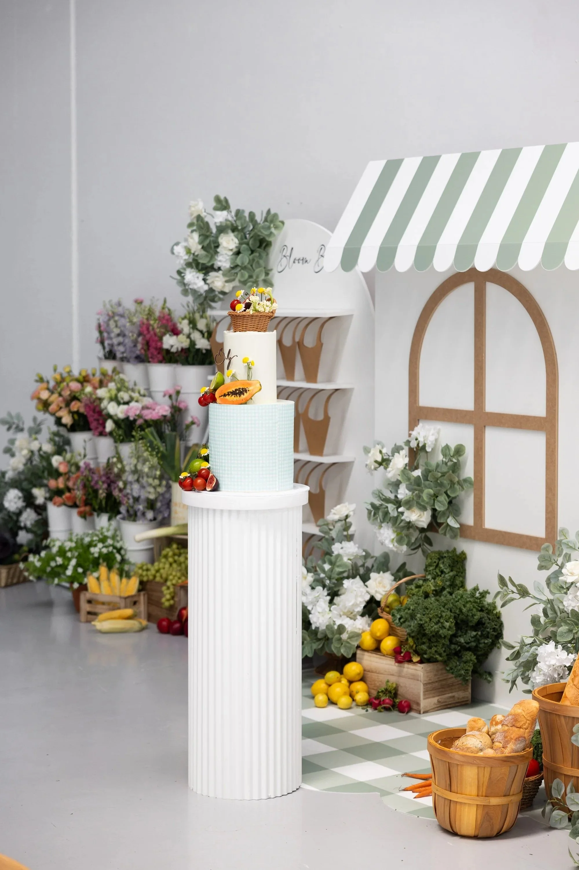 A display of various flowers, fruits, and baked goods arranged in baskets and containers, with a white cake topped with a small basket of berries and flowers, set against a backdrop of a farmers market scene.