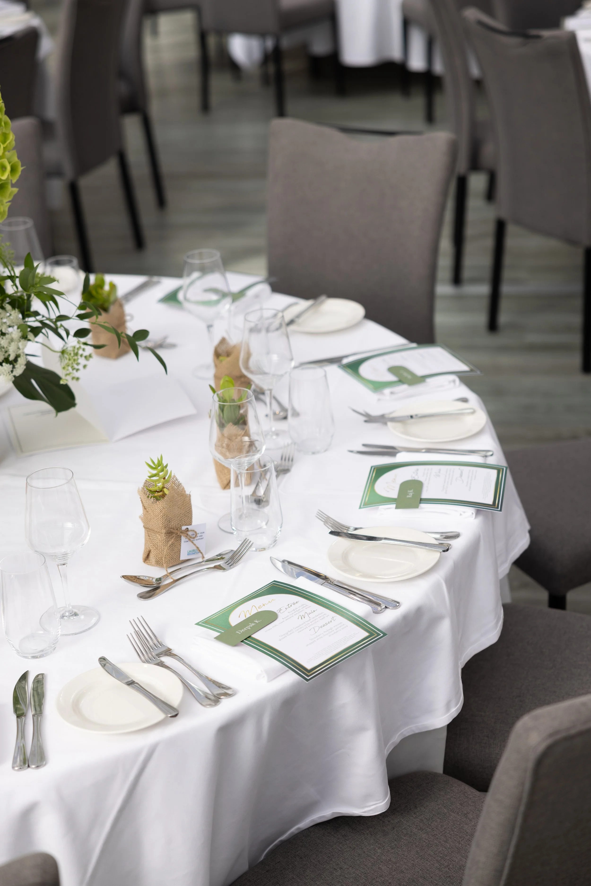 A round banquet table set with white tablecloth, glassware, silverware, menus, small potted succulents wrapped in burlap, and floral centerpiece, arranged for a formal event in a room with gray chairs and wooden flooring.