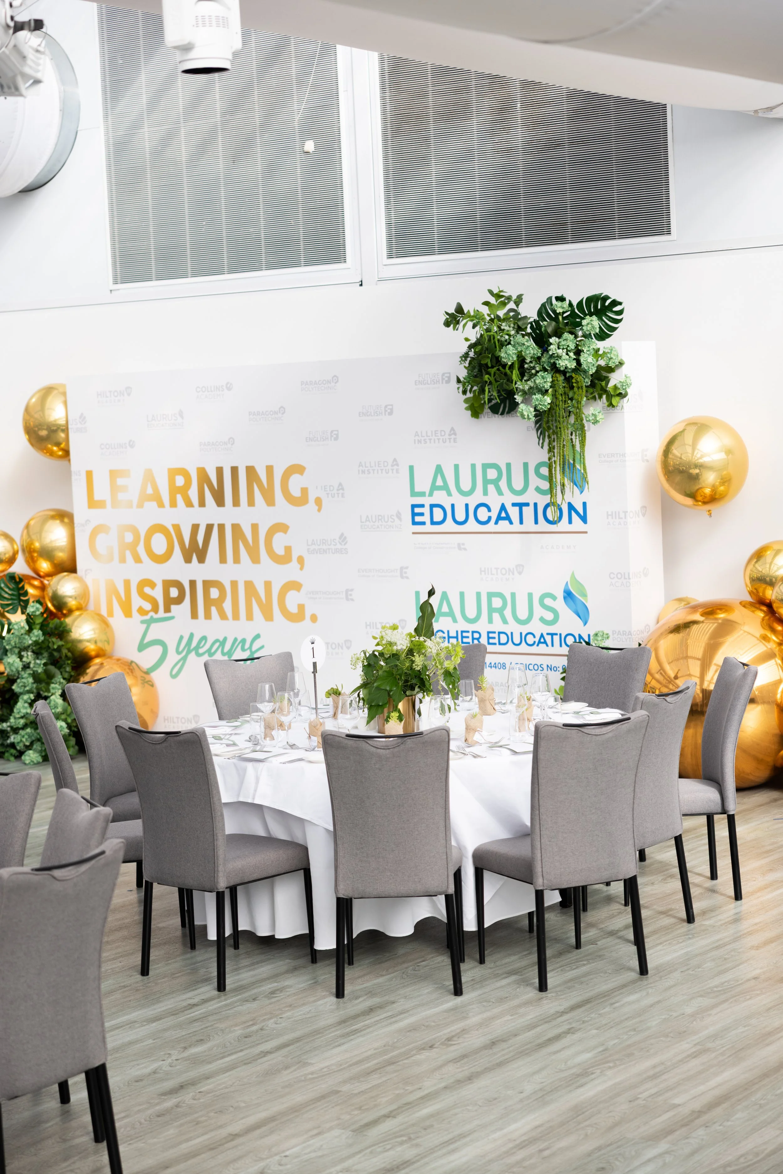 A decorated event space with a round table set with glassware and a centerpiece, gray chairs, a backdrop with logos and motivational words, and gold balloons, in front of a wall with large green floral arrangements and banners celebrating the five-year anniversary of Laurus Education.