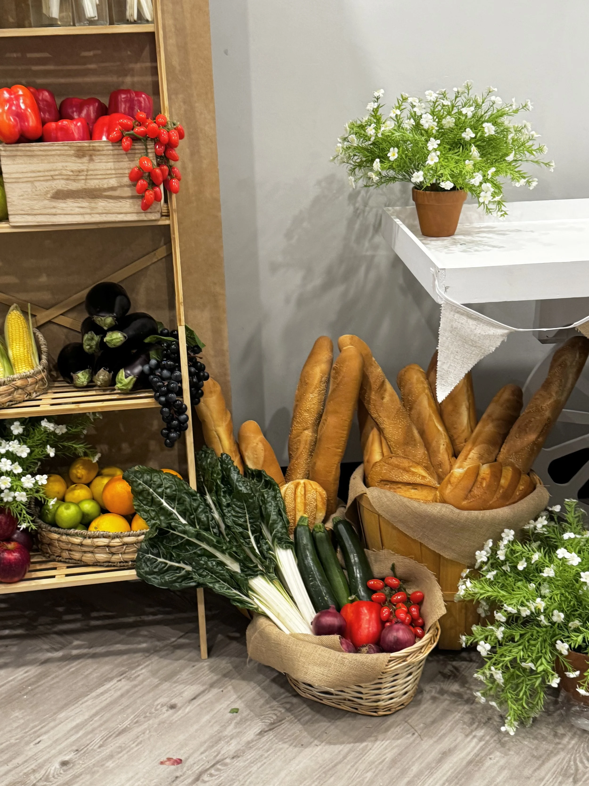 A display of fresh vegetables and flowers, including red bell peppers, cherry tomatoes, eggplants, zucchinis, carrots, onions, and white flowers in pots, arranged on baskets, a white table, and a wooden shelf.