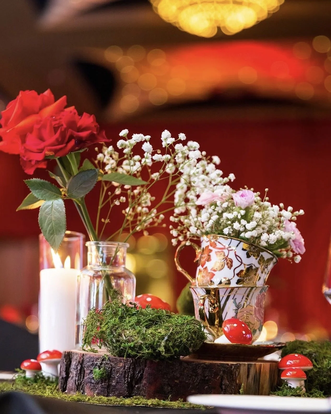 Vase with red roses and white baby’s breath, decorative teacup with pink carnations and baby's breath, moss, red mushrooms, and a lit white candle on a wooden surface, with blurred colorful background lights.