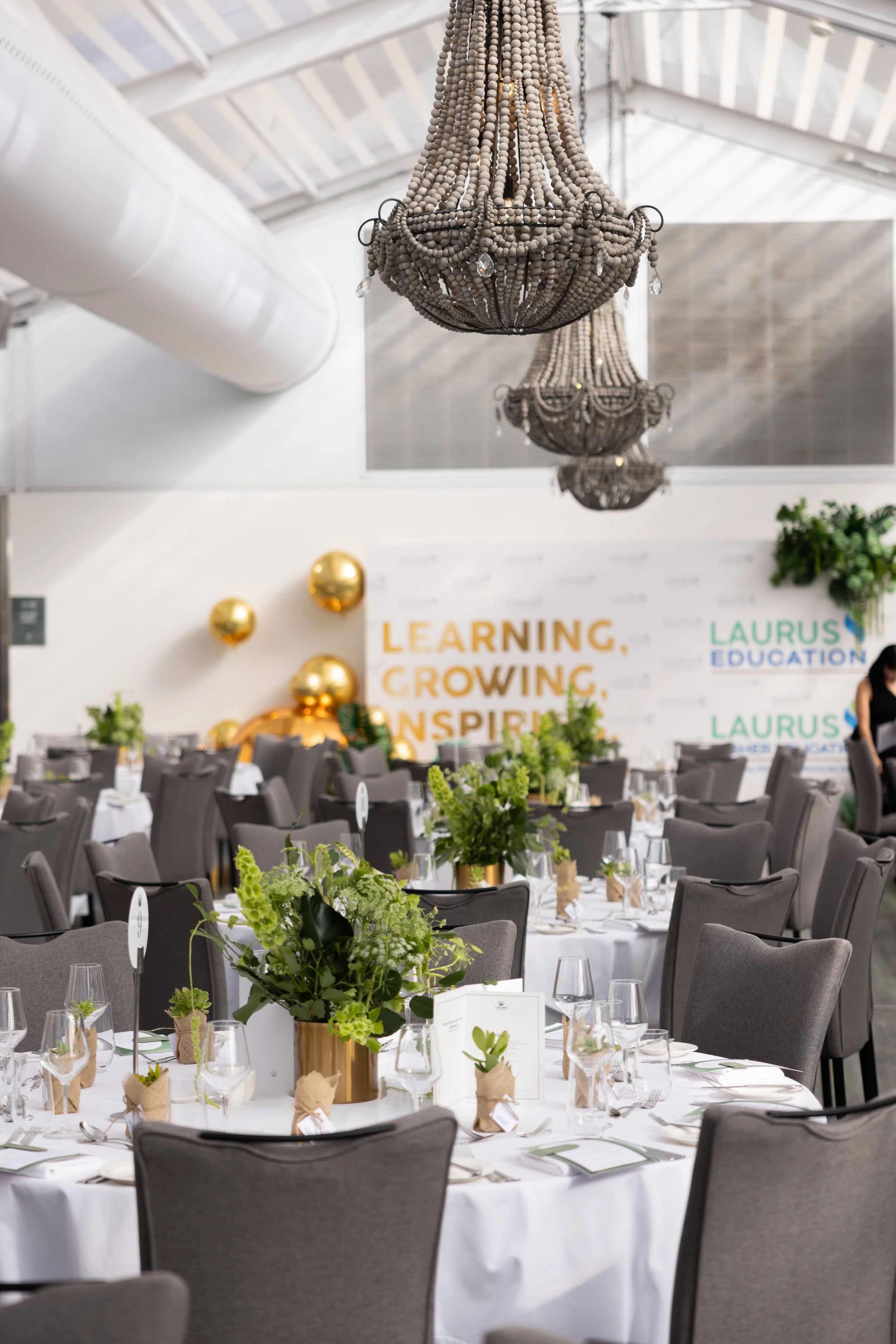 Elegant event space with round tables decorated with green floral centerpieces, set for dining with wine glasses and place cards, featuring large chandeliers hanging from a bright, airy ceiling. The background includes a wall with balloons and a sign that reads 'Learning, Growing, Inspiring' and 'Laurus Education'.