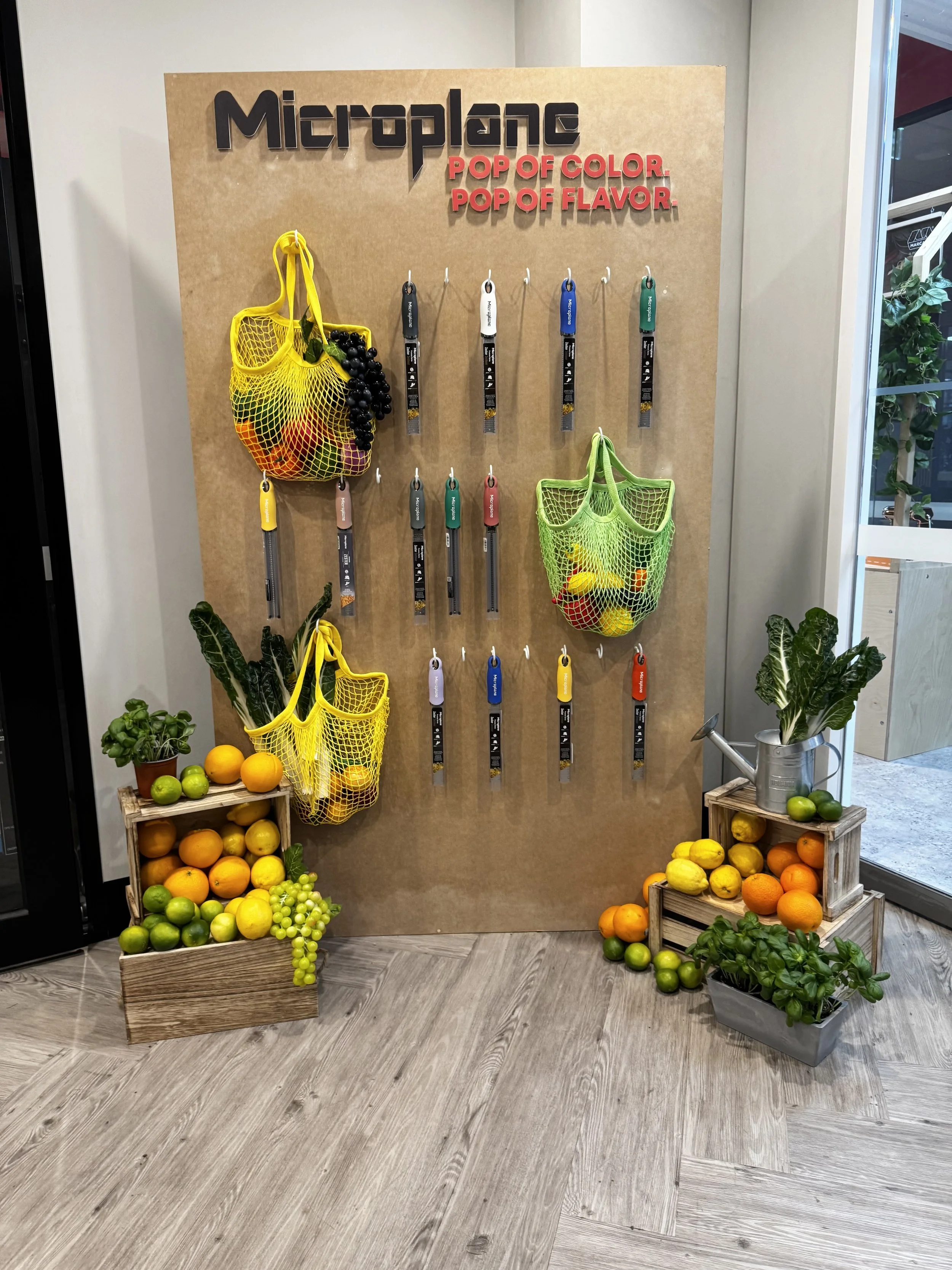 Display of colorful rubber handle garden tools hung on a wooden board, with baskets of fresh citrus fruits and green plants arranged on crates and the floor around it.