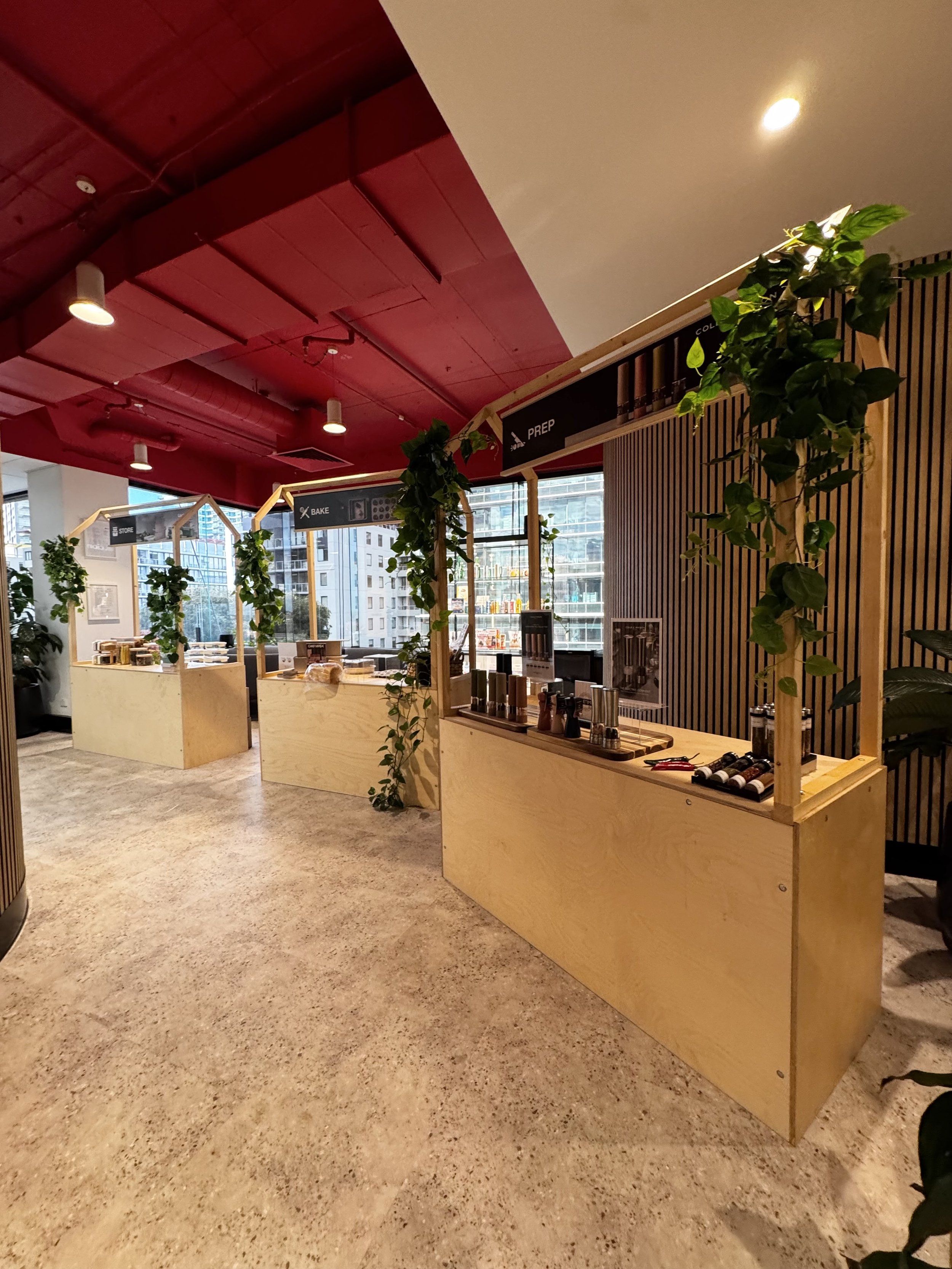 Interior of a modern office or cafeteria with wooden decor, green plants, and food service counters, large windows showing city buildings outside, ceiling painted red with ceiling lights.