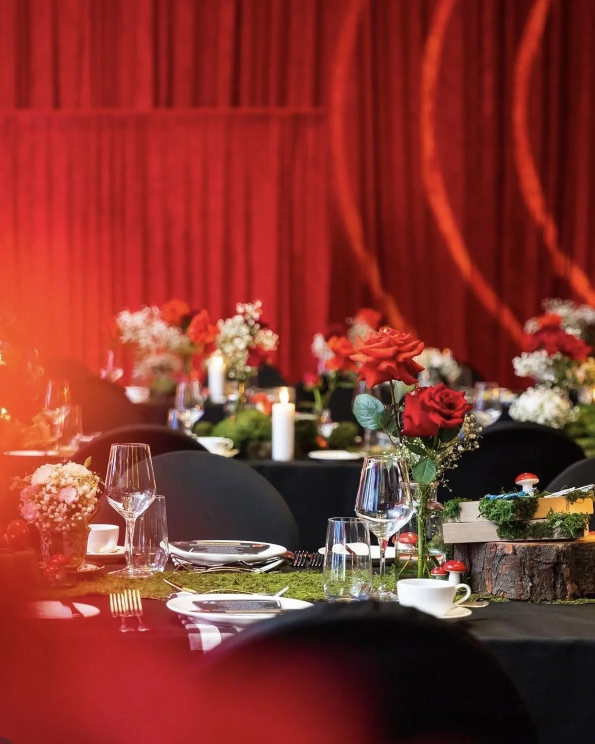Elegant event table setup with floral centerpieces featuring red roses and small white flowers, glassware, plates, and candles, against a red curtain backdrop.