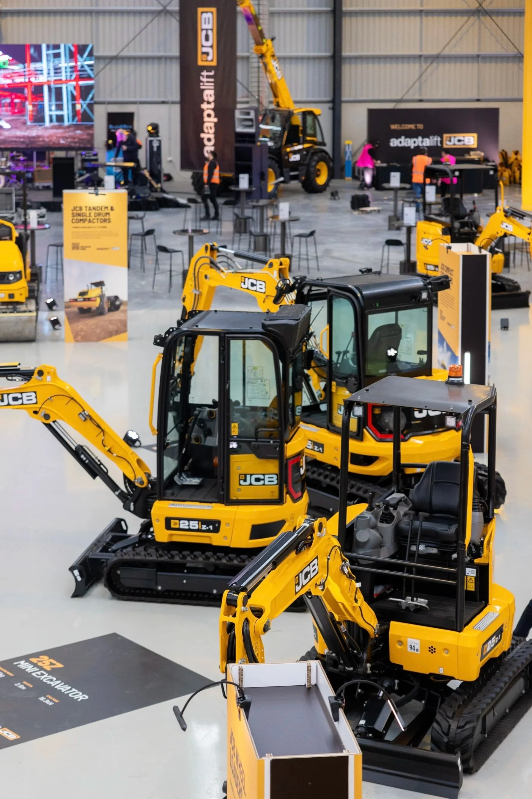 Display of yellow JCB compact excavators at an indoor trade show with banners and informational signs.