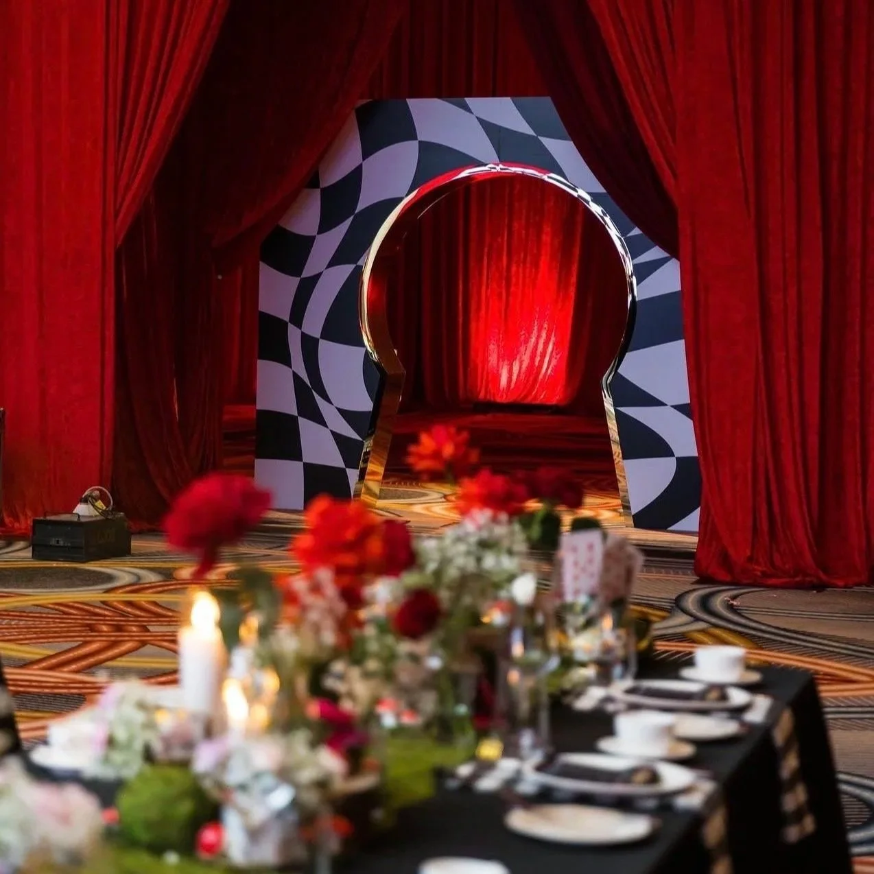 A decorated event space with a stage opening framed by red curtains. The stage has a black and white patterned archway, and in the foreground, there is a table with floral centerpieces and place settings, with a blurred background of flowers and candles.