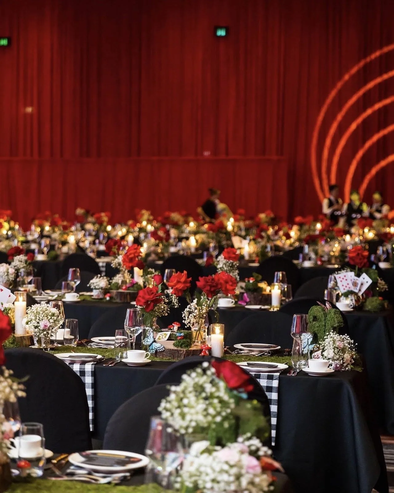 Elegant banquet hall decorated with red, white, and pink flowers, candles, and candles with black tablecloths and black chairs. Red backdrop with orange light arcs, and waitstaff in black and white uniforms.