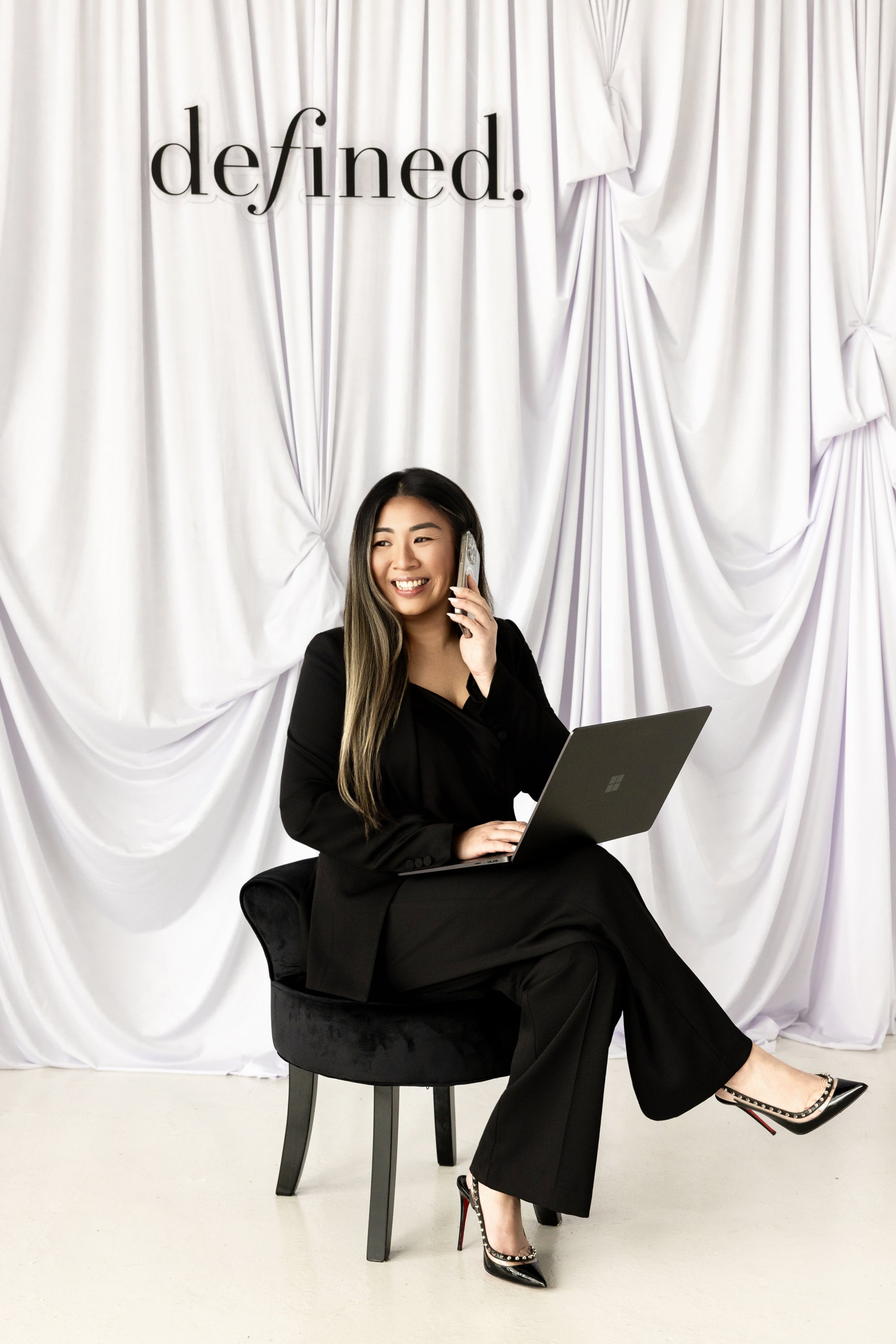 A woman in a black suit sitting on a small stool, talking on a cell phone, with a laptop on her lap, in front of a white draped backdrop with the word 'defined.' written on it.