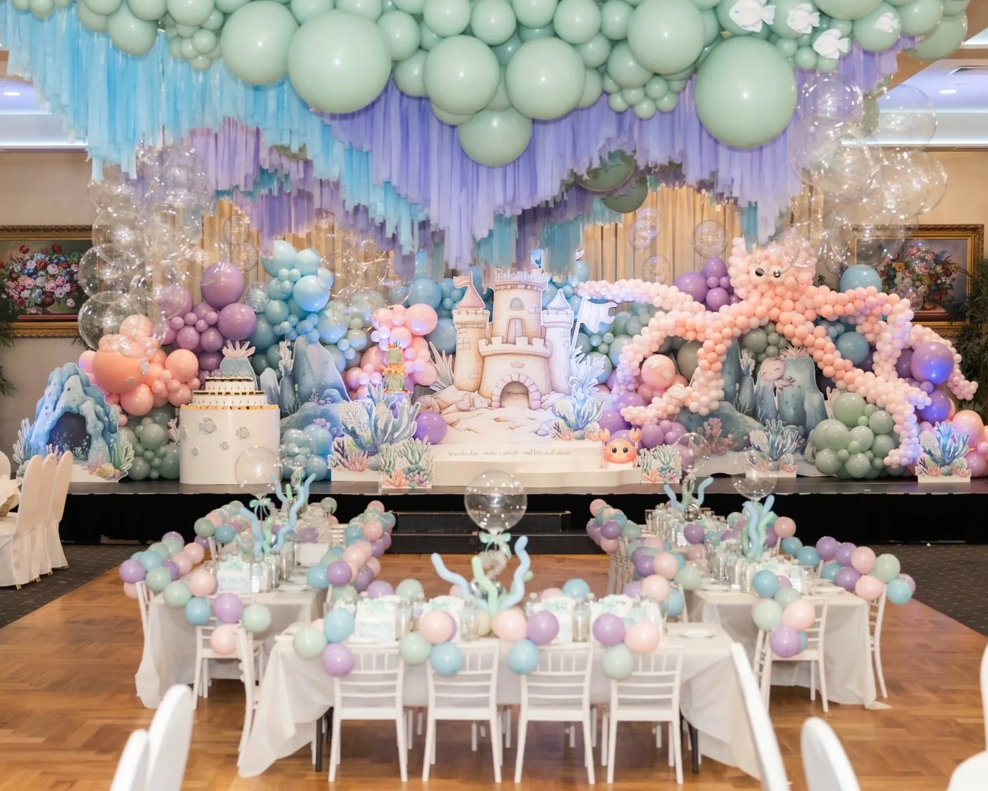 A decorated indoor space for a celebration features an abundance of pastel-colored balloons in shades of green, pink, purple, and blue, creating a canopy overhead and surrounding a whimsical castle-themed centerpiece on a table. The scene includes decorative elements like faux coral, sea creatures, and an oversized pink octopus made of balloons, with tables set with white chairs and tableware beneath the balloon decorations.