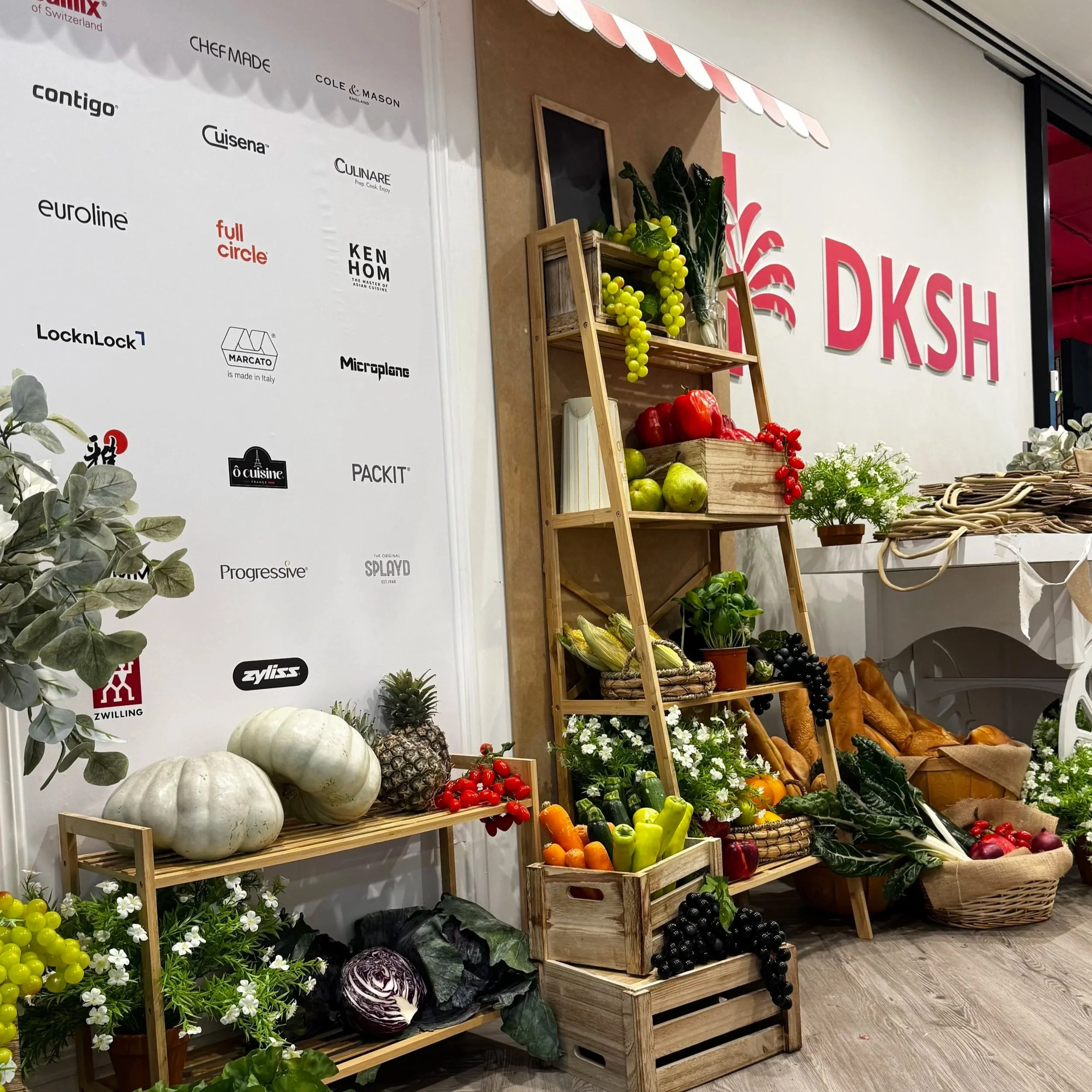 Display of fresh vegetables, fruits, and plants on wooden shelves at an indoor market or store. There are pumpkins, grapes, tomatoes, carrots, bell peppers, radishes, eggplants, leafy greens, and decorative plants, with a branded backdrop on the wall.