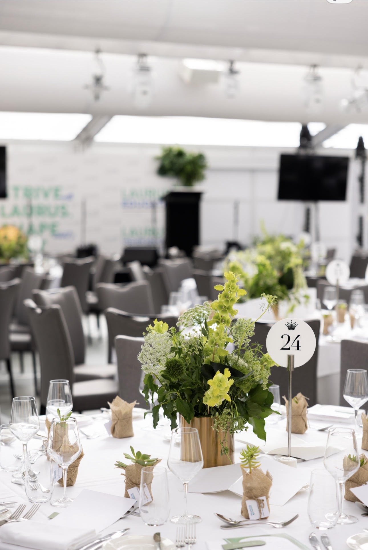 A banquet table set with glassware, napkins, and a floral centerpiece in a gold vase at an event venue. The table has a sign with the number 24. In the background, there are numerous chairs and a blurred view of a large room with a high ceiling and electronic screens.