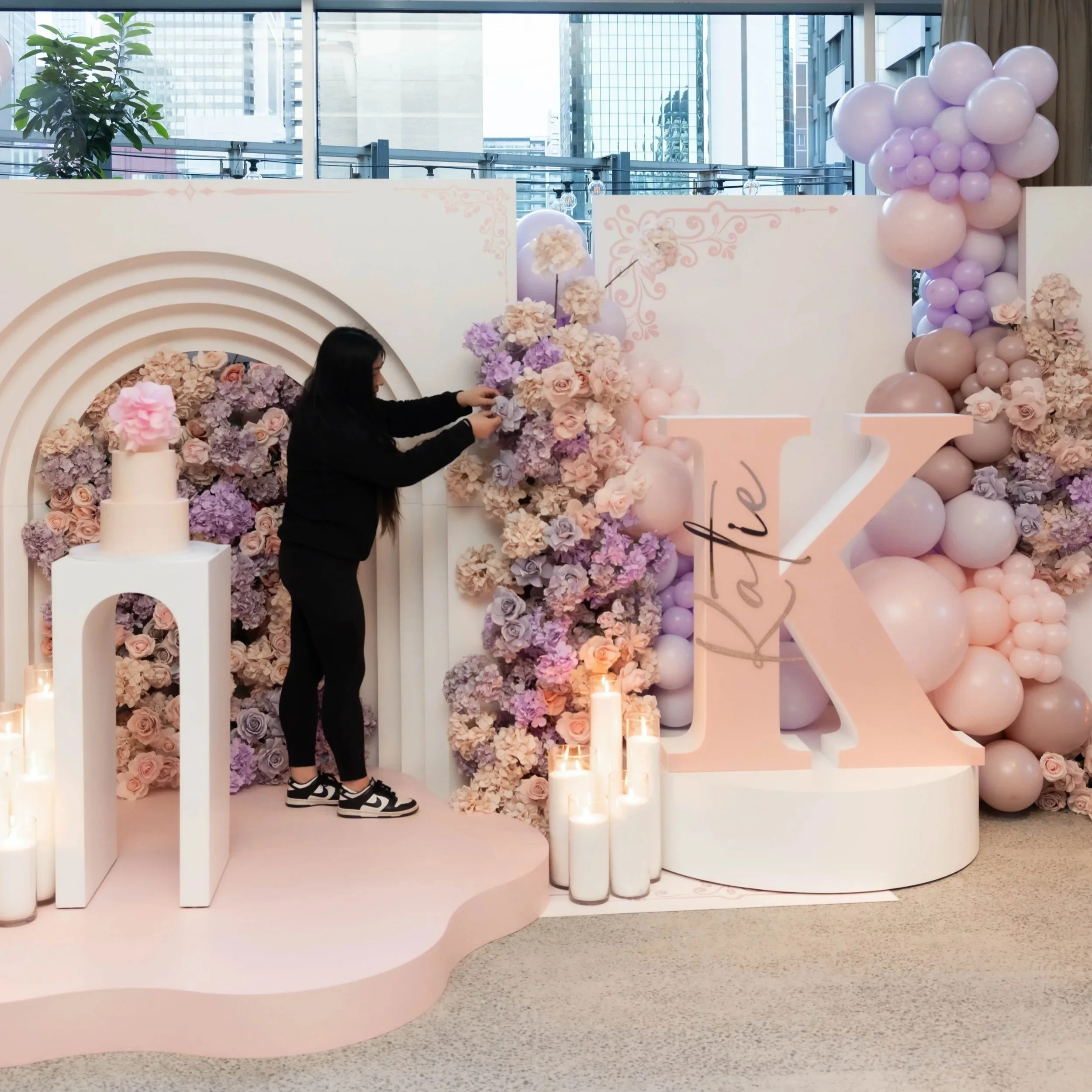 Behind-the-scenes event styling featuring pastel pink and purple floral arrangements, balloon decorations and a custom event backdrop.