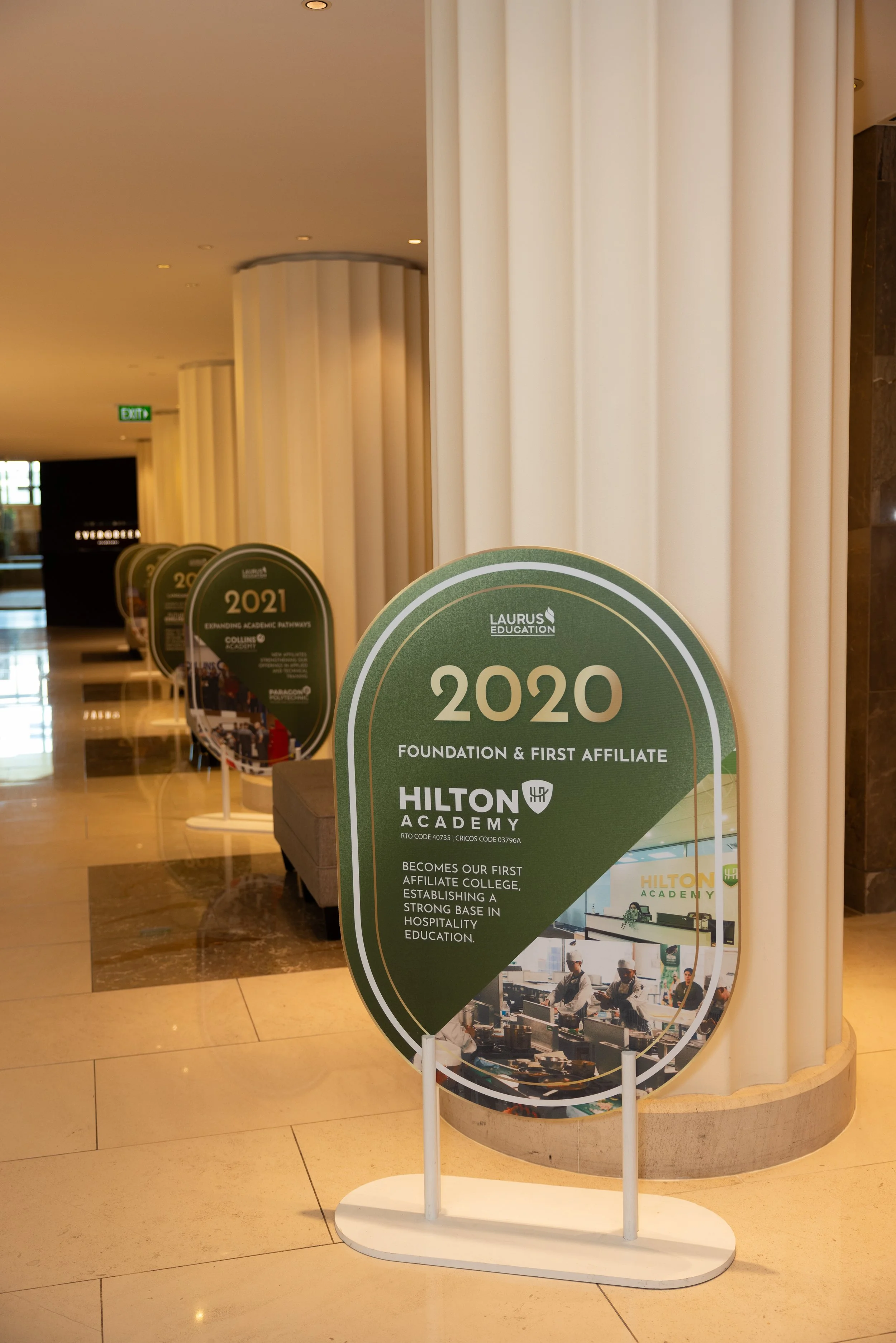 Lobby area with vertical white curtains, informational signs on stands, including one for Hilton Academy 2020, and an exit sign in the background.