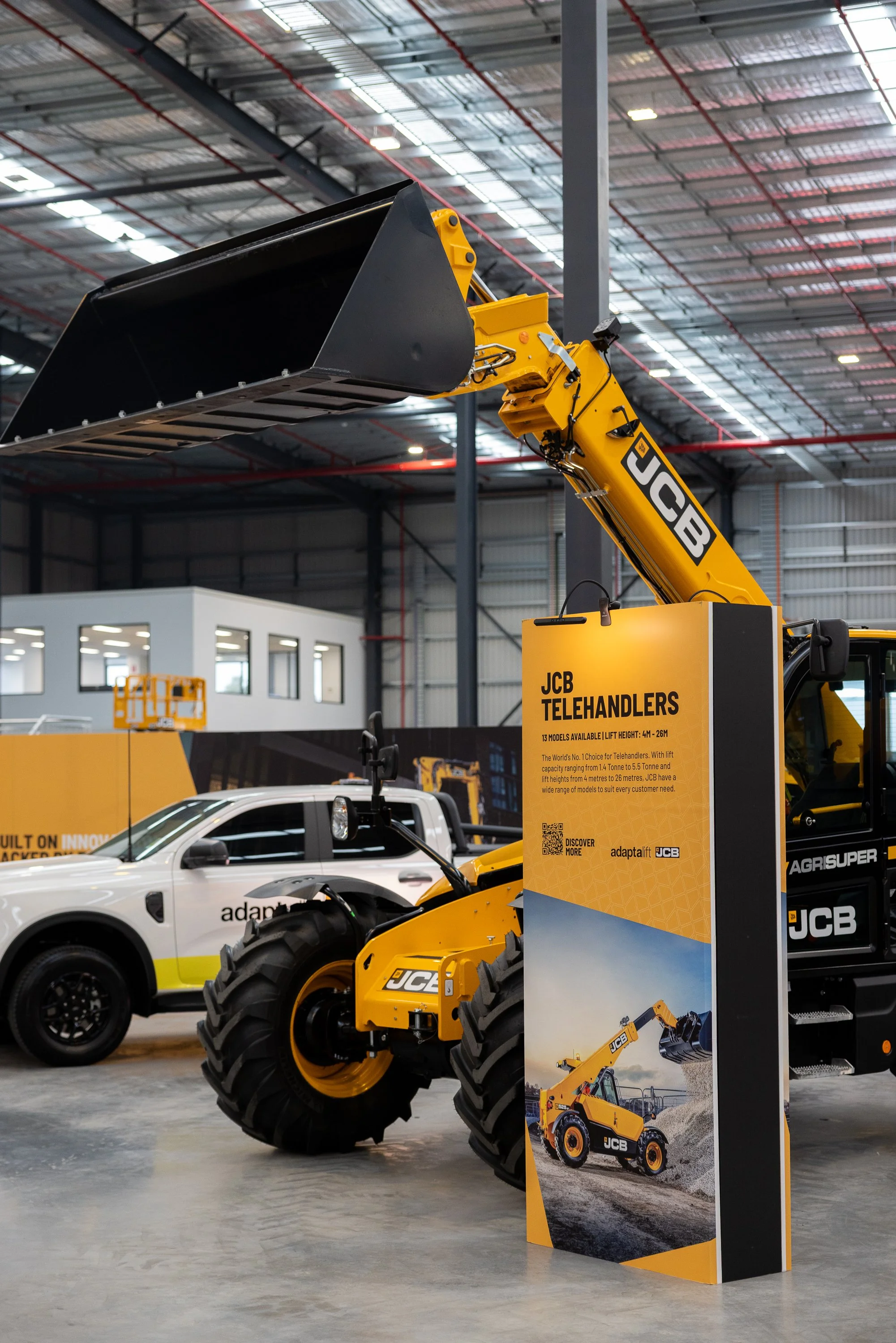 Indoor display of construction equipment, including a yellow JCB telehandler and a white vehicle with black wheels, inside a large warehouse or exhibition hall with high ceiling and metallic beams.