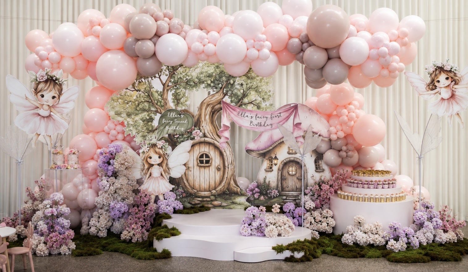 Decorative fairy tale-themed birthday party setup with pink and purple balloons, fairy figurines, flower arrangements, and a cake on a white cake stand. Background features illustrated fairy houses and trees.