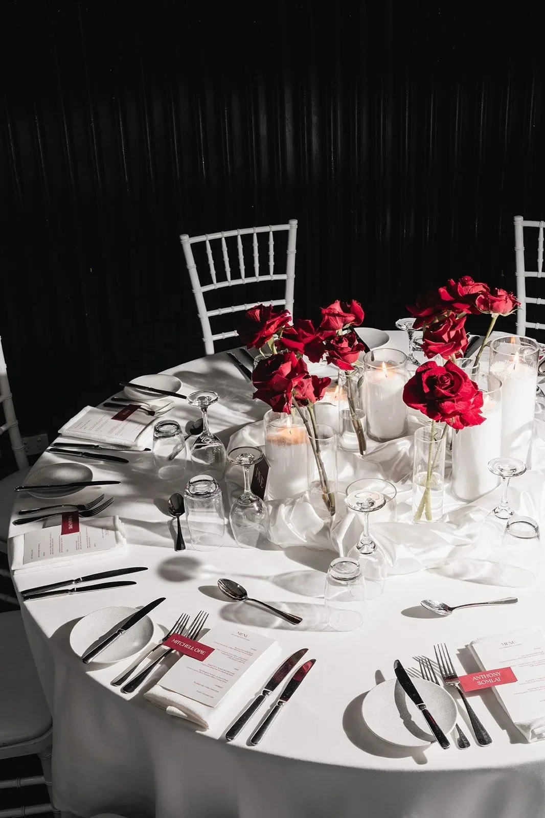 Round table set for a formal event with white tablecloth, red flowers in tall vases, candles, glassware, and silverware, placed in front of white chairs with a black wall background