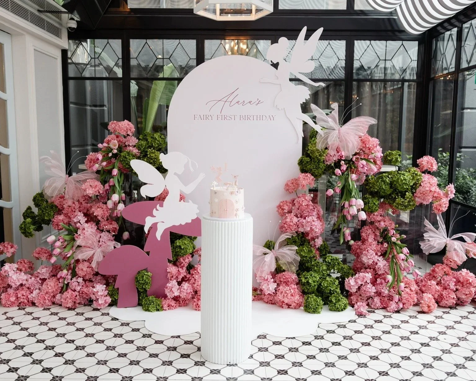 Decorative backdrop for a child's fairy-themed birthday party with pink and green flowers, fairy silhouettes, and a white cake on a stand in front.