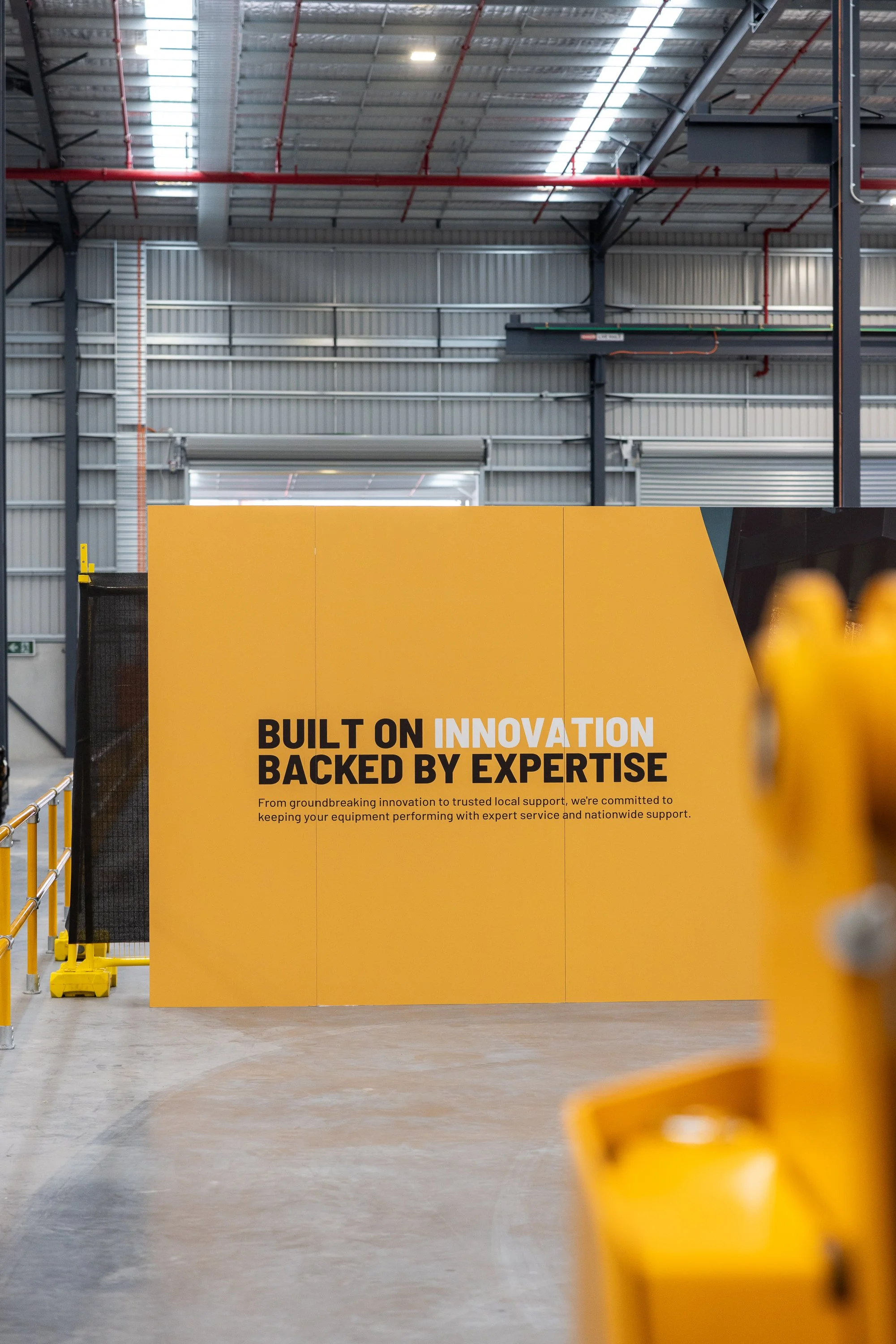 Industrial warehouse interior with a yellow sign that reads, 'Built on Innovation Backed by Expertise.' The background features metal walls, a large roller door, and a high ceiling with red pipes.