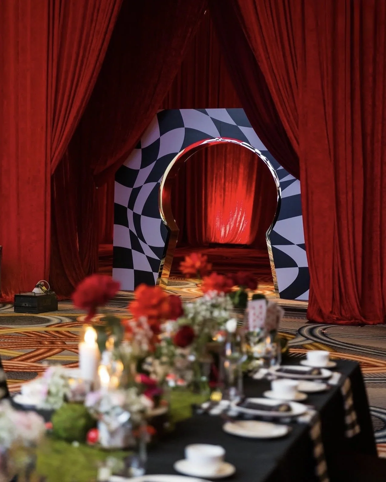 A decorated event space with a stage framed by red velvet curtains, a circular doorway with black and white geometric patterns, and floral centerpieces on a banquet table in the foreground.