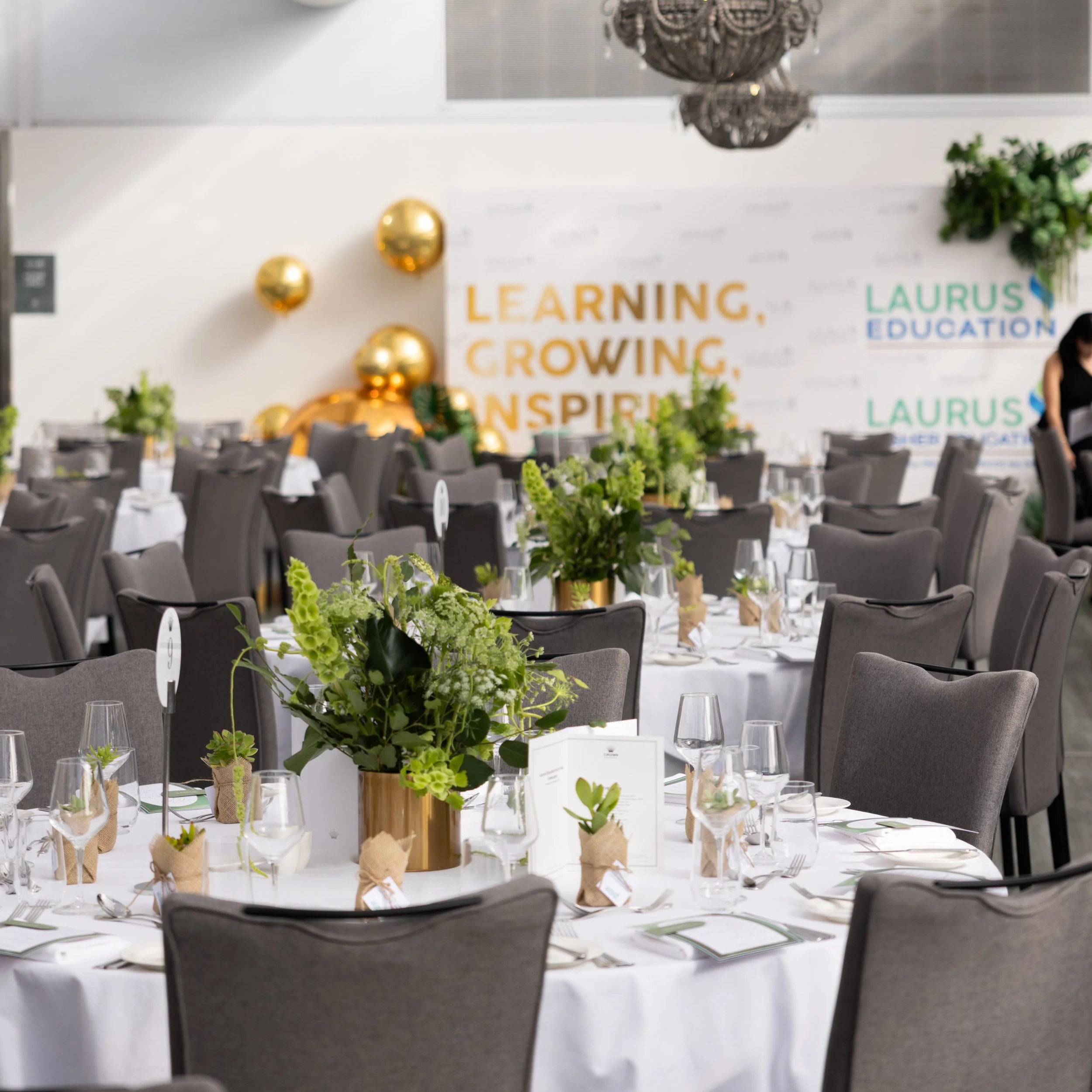 Elegant event space with round tables covered in white tablecloths, decorated with green floral centerpieces wrapped in burlap, and surrounded by gray chairs. A large wall prompt reads, "Learning, Growing, Inspiring." The background features a banner displaying the logo 'Laurus Education' and some decorative elements including gold balloons and a chandelier.