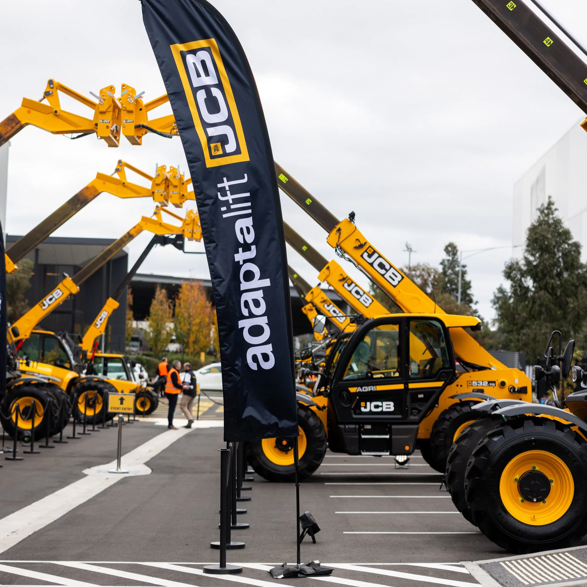 Outdoor display of yellow construction equipment with JCB branding, arranged in rows with event signage, banners, and a cloudy sky overhead.