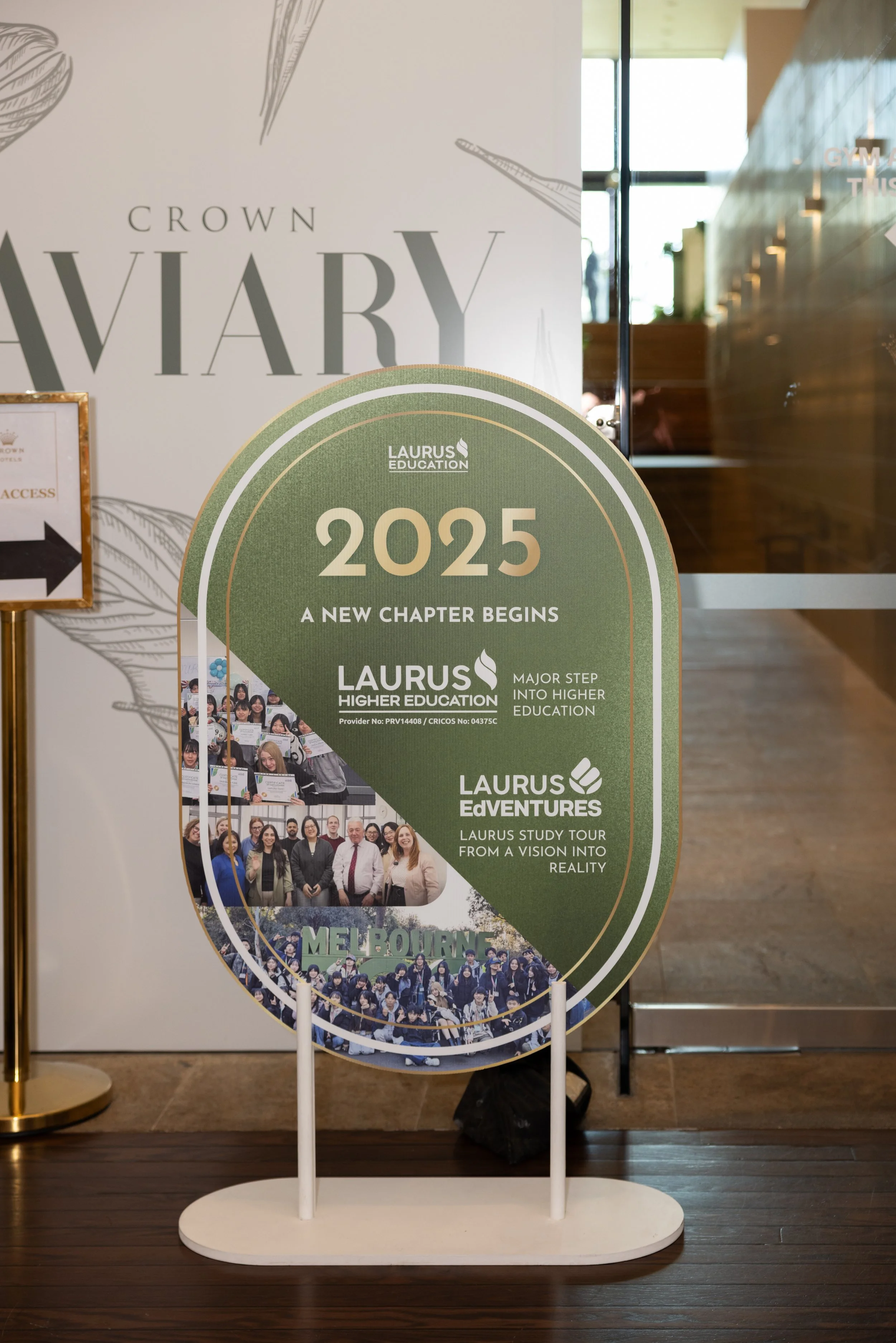 A vertical display board with a large sign reading 'CROWN UNIVERSITY' at the top, featuring a graphic design of feathers. Below this sign, there are multiple smaller images and text promoting Laurus Education, with a focus on the year 2025, and phrases like 'A NEW CHAPTER BEGINS' and 'MAJOR STEP INTO HIGHER EDUCATION.' The display is positioned on a white circular stand inside a building with wooden floors and glass doors.