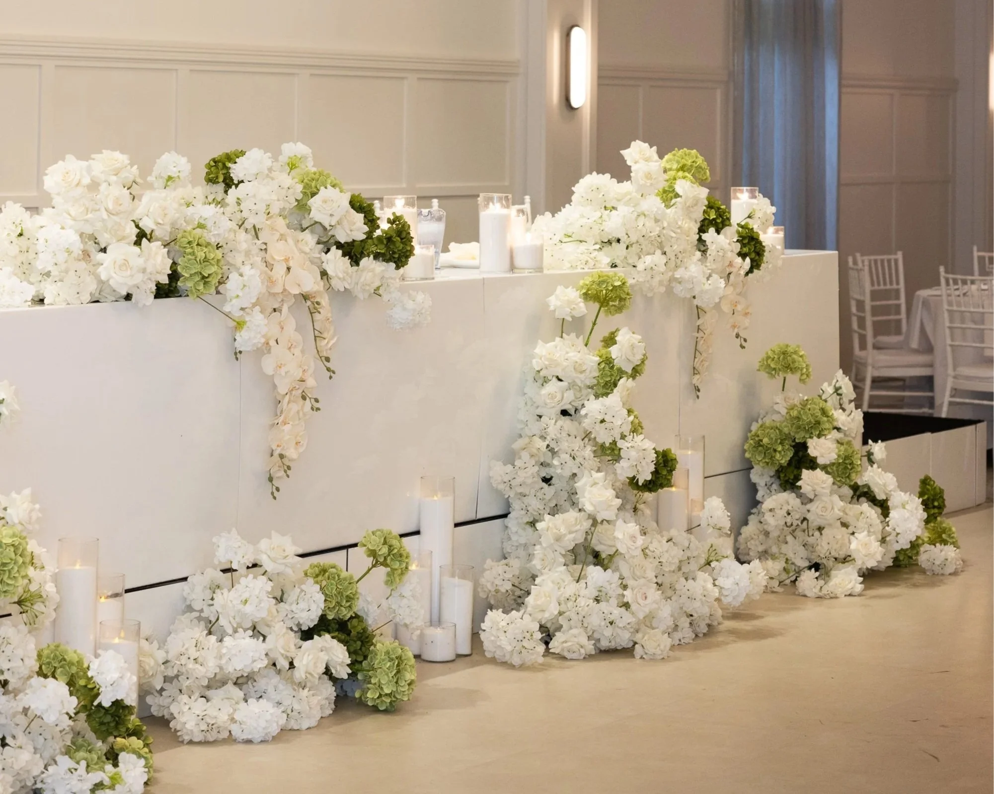 Elegant white floral wedding or event decor featuring white roses, hydrangeas, and green accents, with scattered candles in glass holders on a white wall backdrop.
