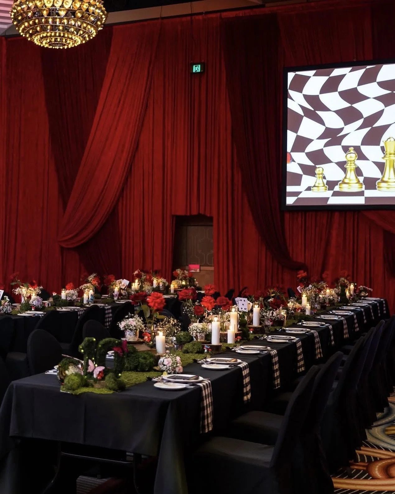 Elegant banquet hall decorated with black tablecloths, red and white floral centerpieces, candles, and black chairs, with a large red curtain backdrop and a chandelier overhead.