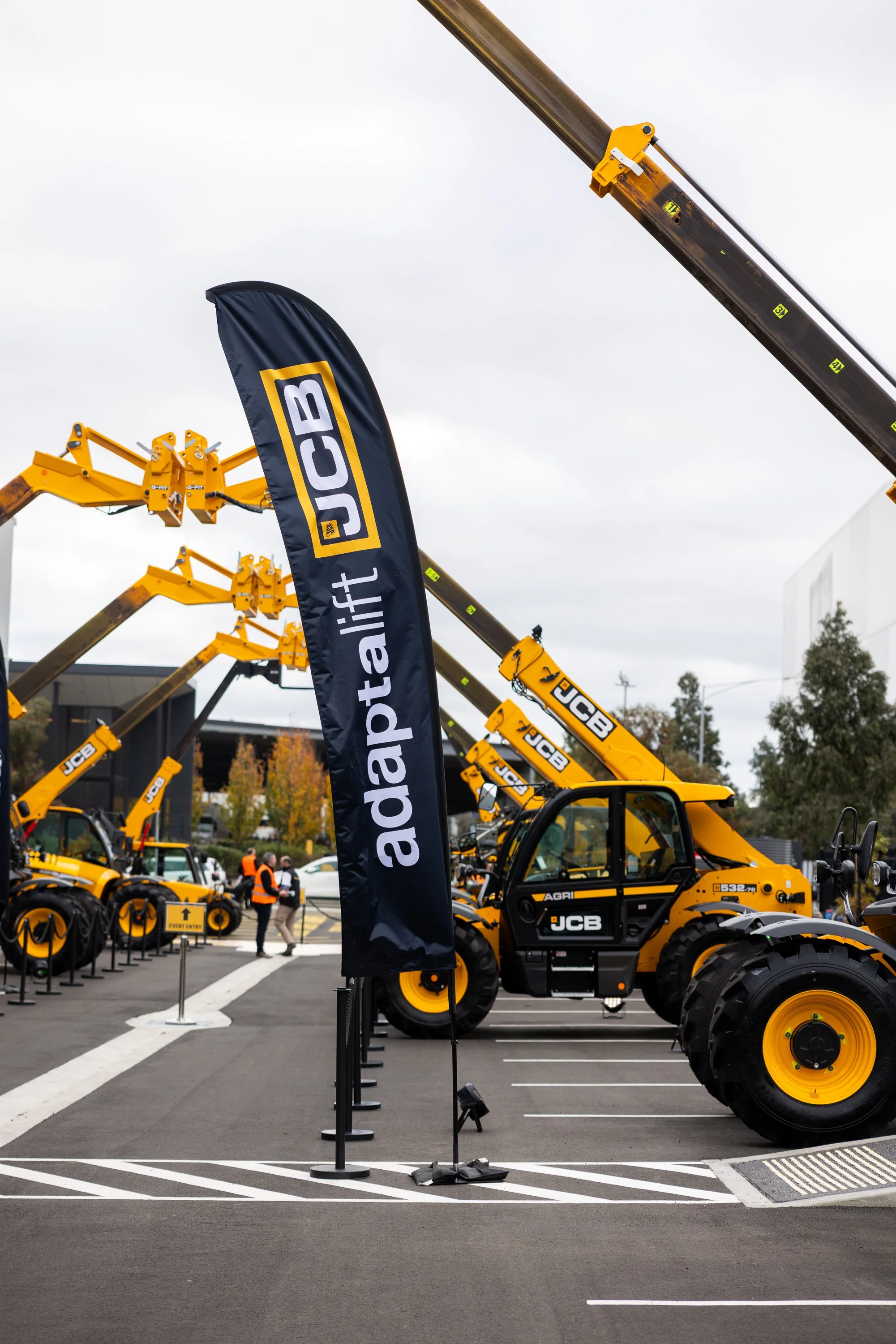 Line of yellow JCB construction equipment on display at an outdoor event with a black flag bearing the JCB logo, overcast sky, and people walking in the background.
