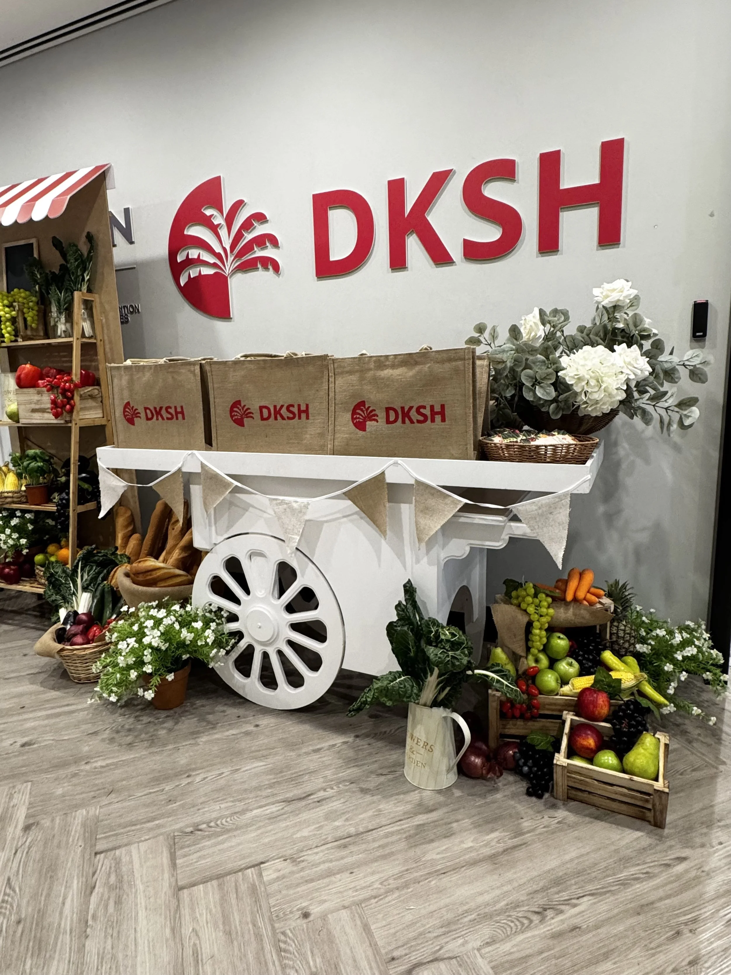 Display of fresh fruits and vegetables arranged on a white cart and surrounding shelves, with a large DKS H logo on the wall behind and decorative flowers.
