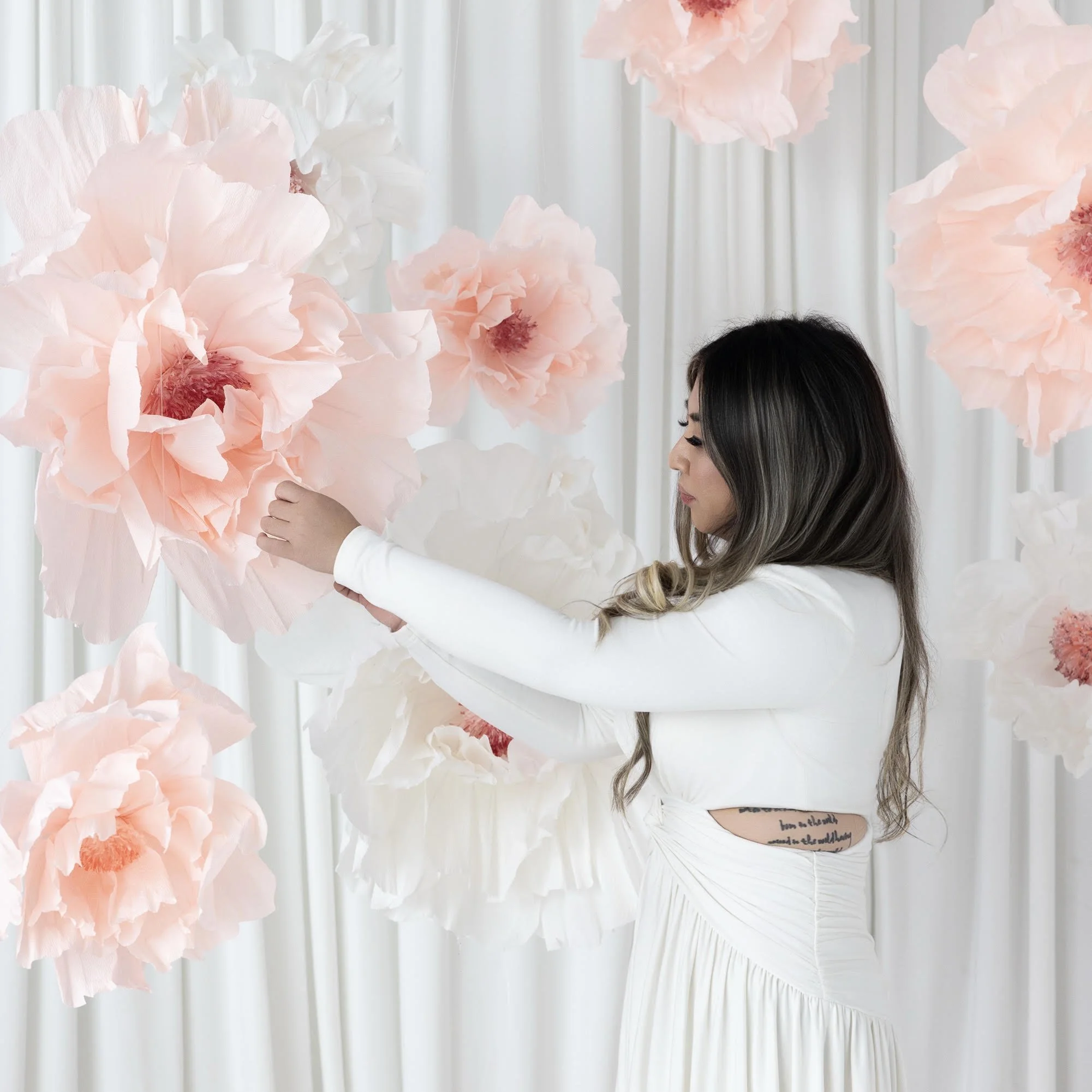 Event stylist arranging oversized blush pink and white paper flowers against a white draped backdrop for a custom floral installation.