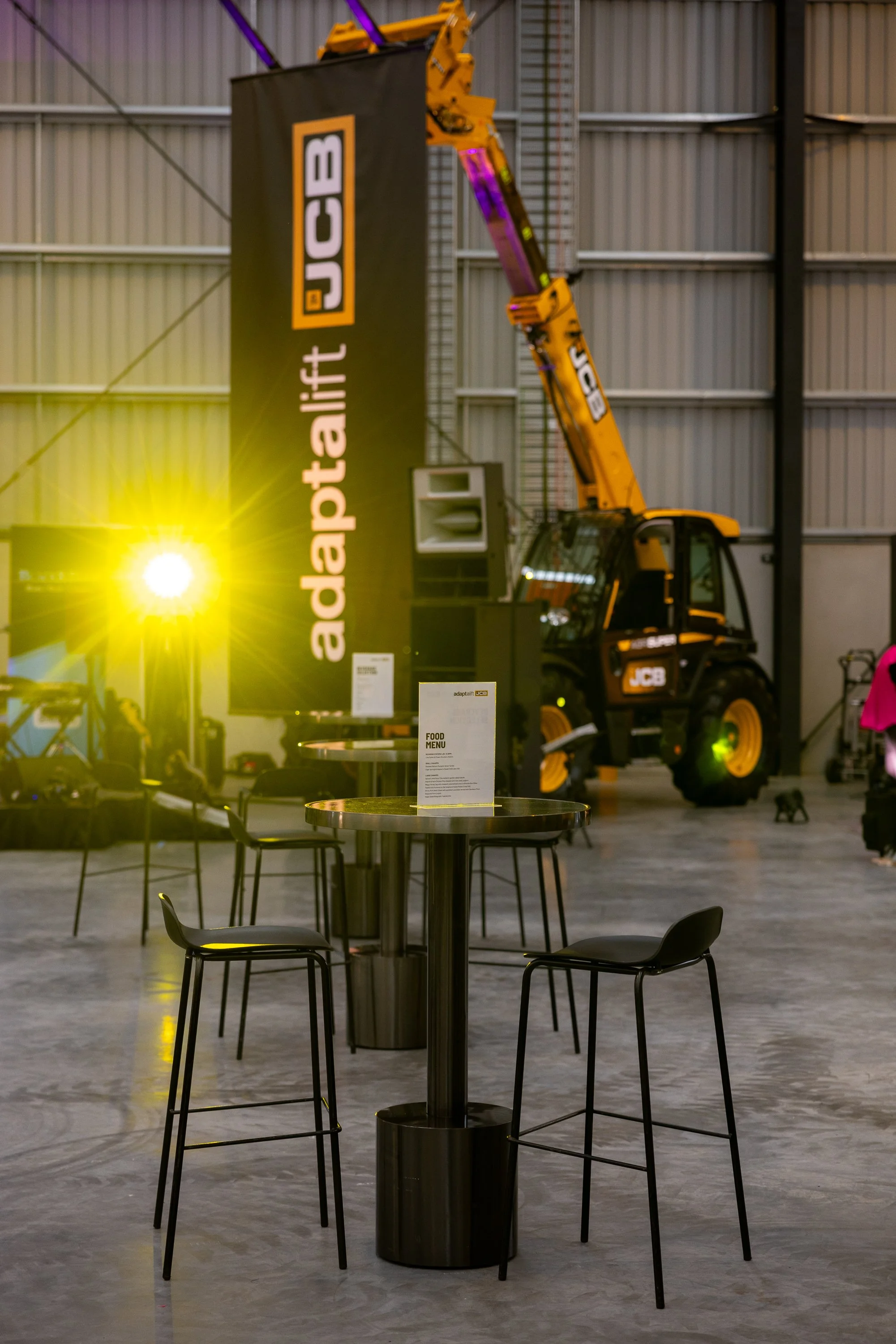 Indoor event space with black tables and chairs, a yellow JCB crane lifting a large advertising banner, and bright yellow and purple lighting.