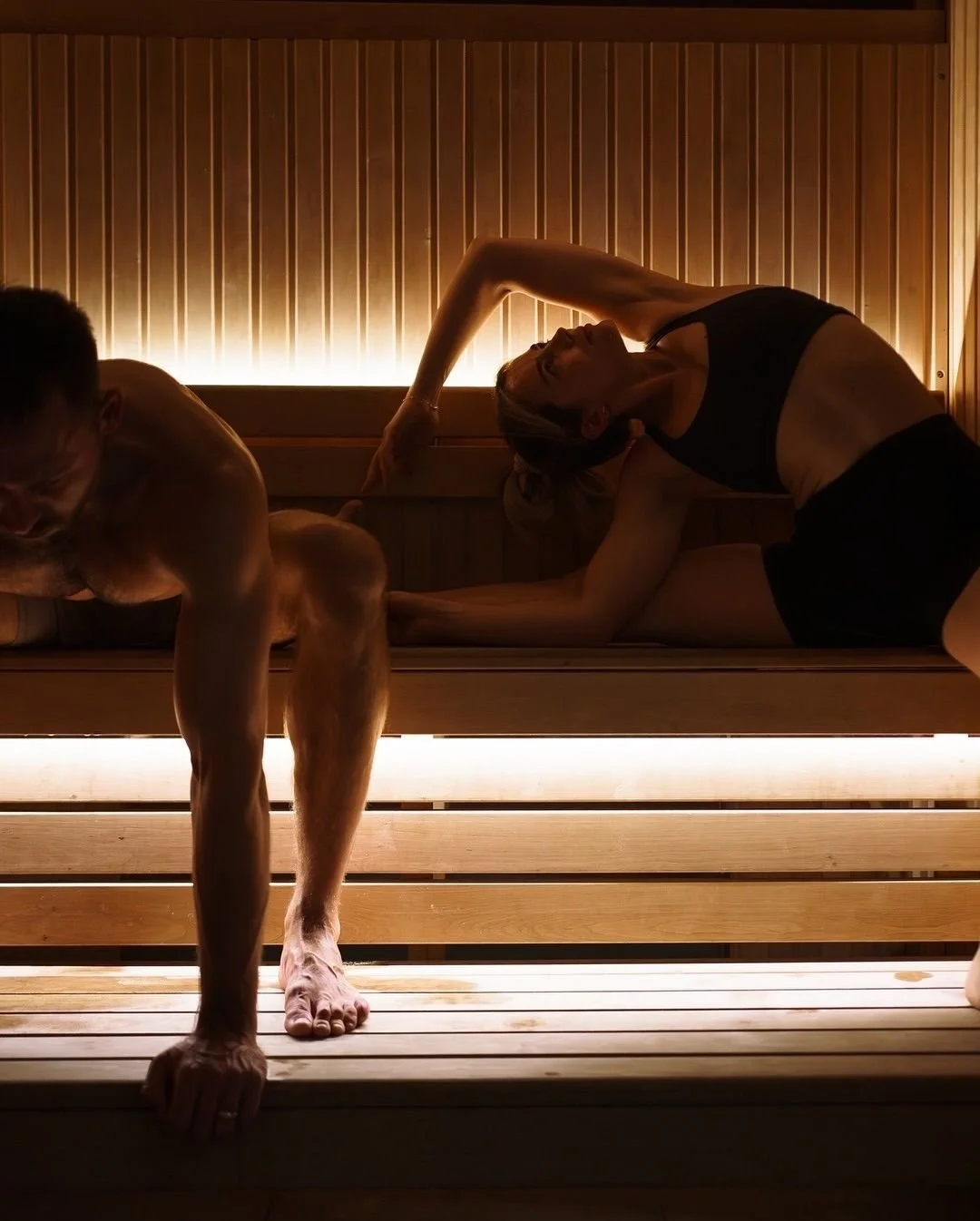 A man and a woman practicing yoga or stretching inside a sauna, with warm lighting and wooden interior.