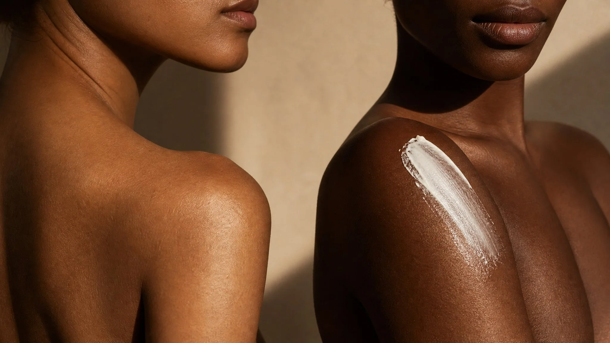 Two women with dark skin, one with light makeup and the other with moisturizer or sunscreen on shoulder.